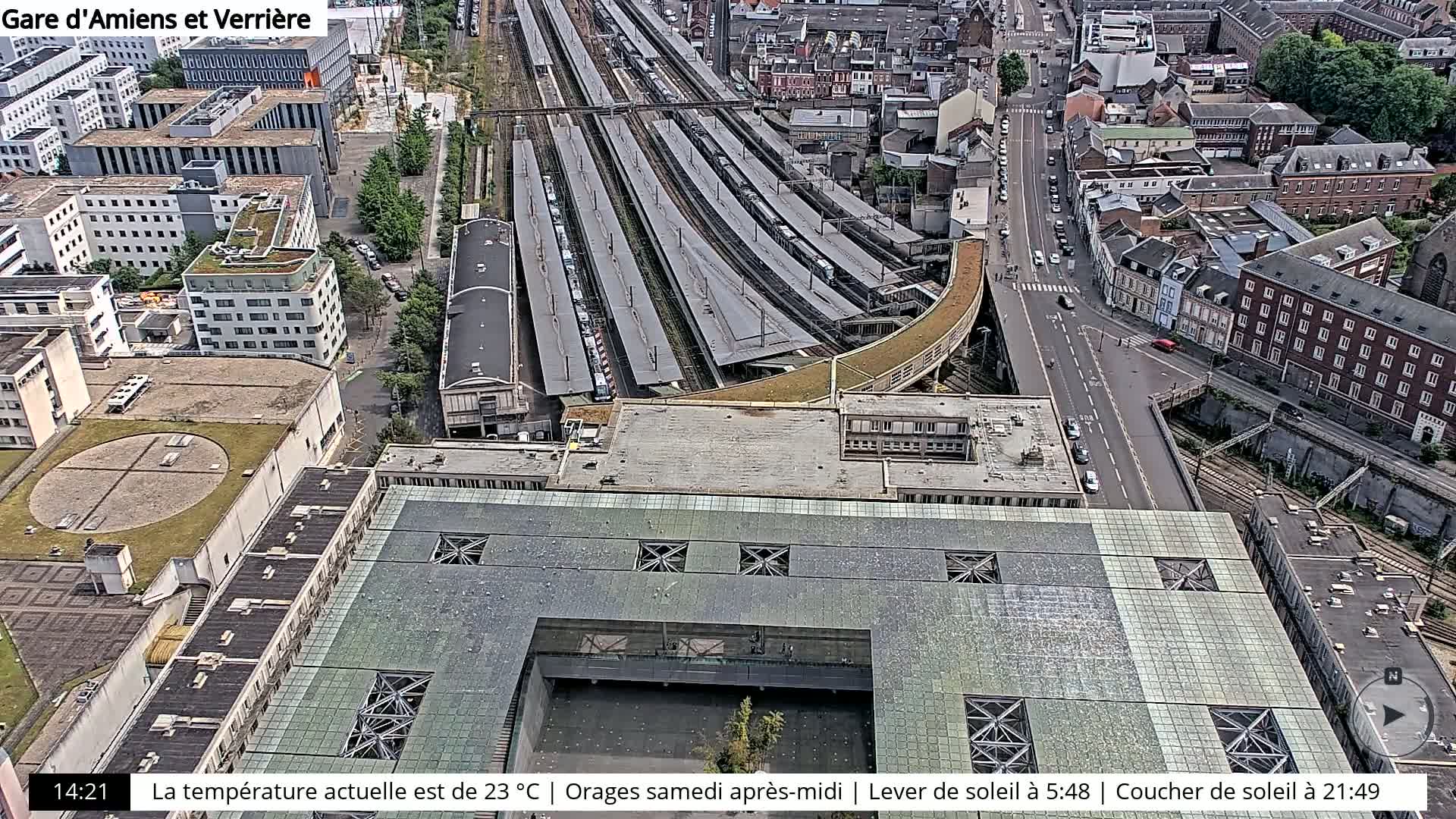 An aerial view of a train station and surrounding city, showing a partly sunny day with a partially green rooftop structure over a courtyard.