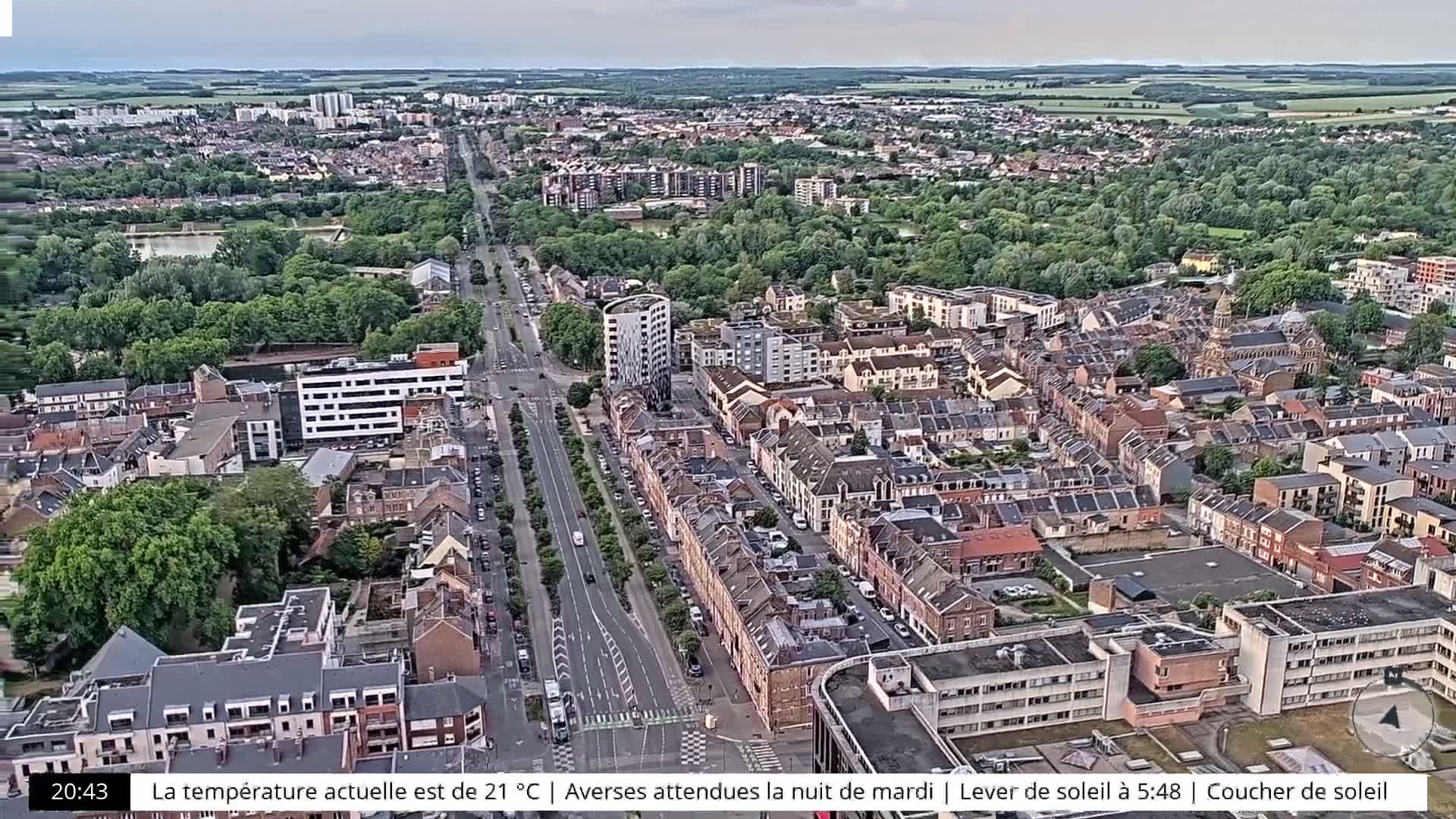 An aerial view of a city with a long road lined with trees and buildings on either side under an overcast sky.