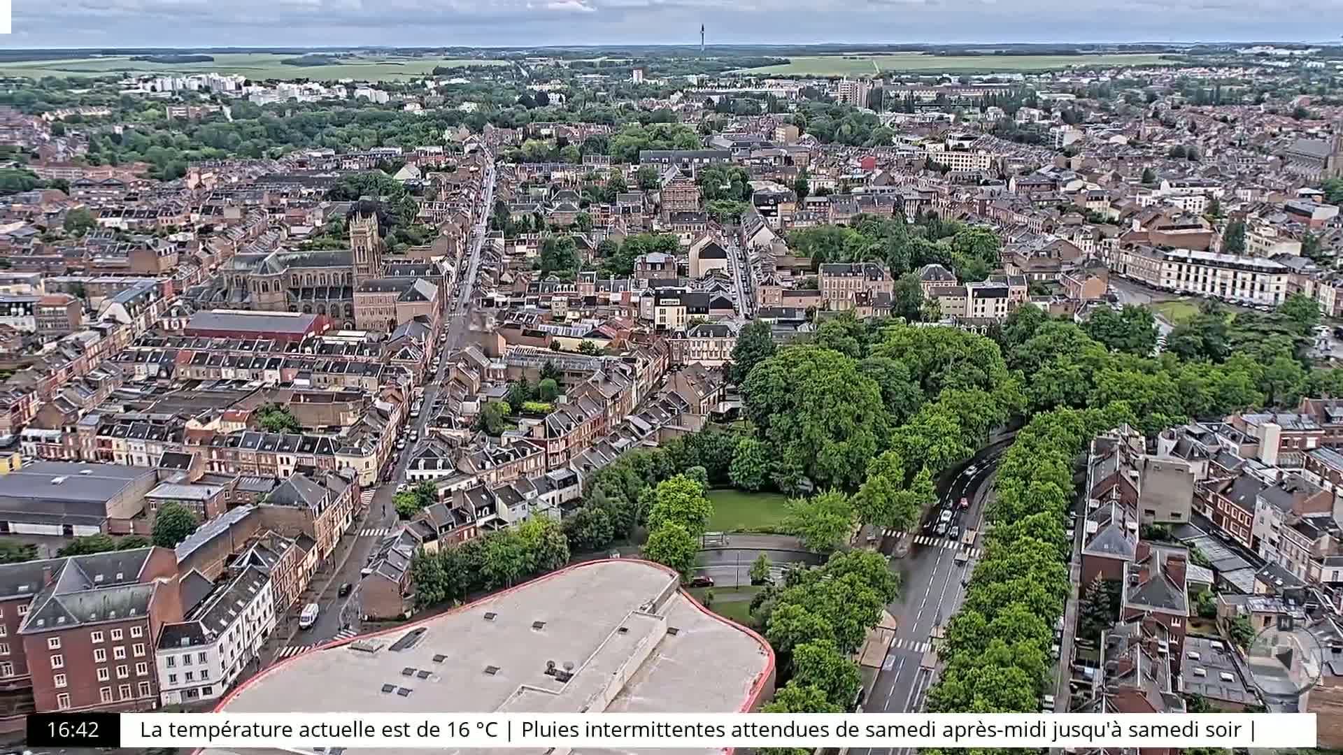 An aerial view shows a densely populated European town with overcast skies, a large green park, and roads winding through the buildings.