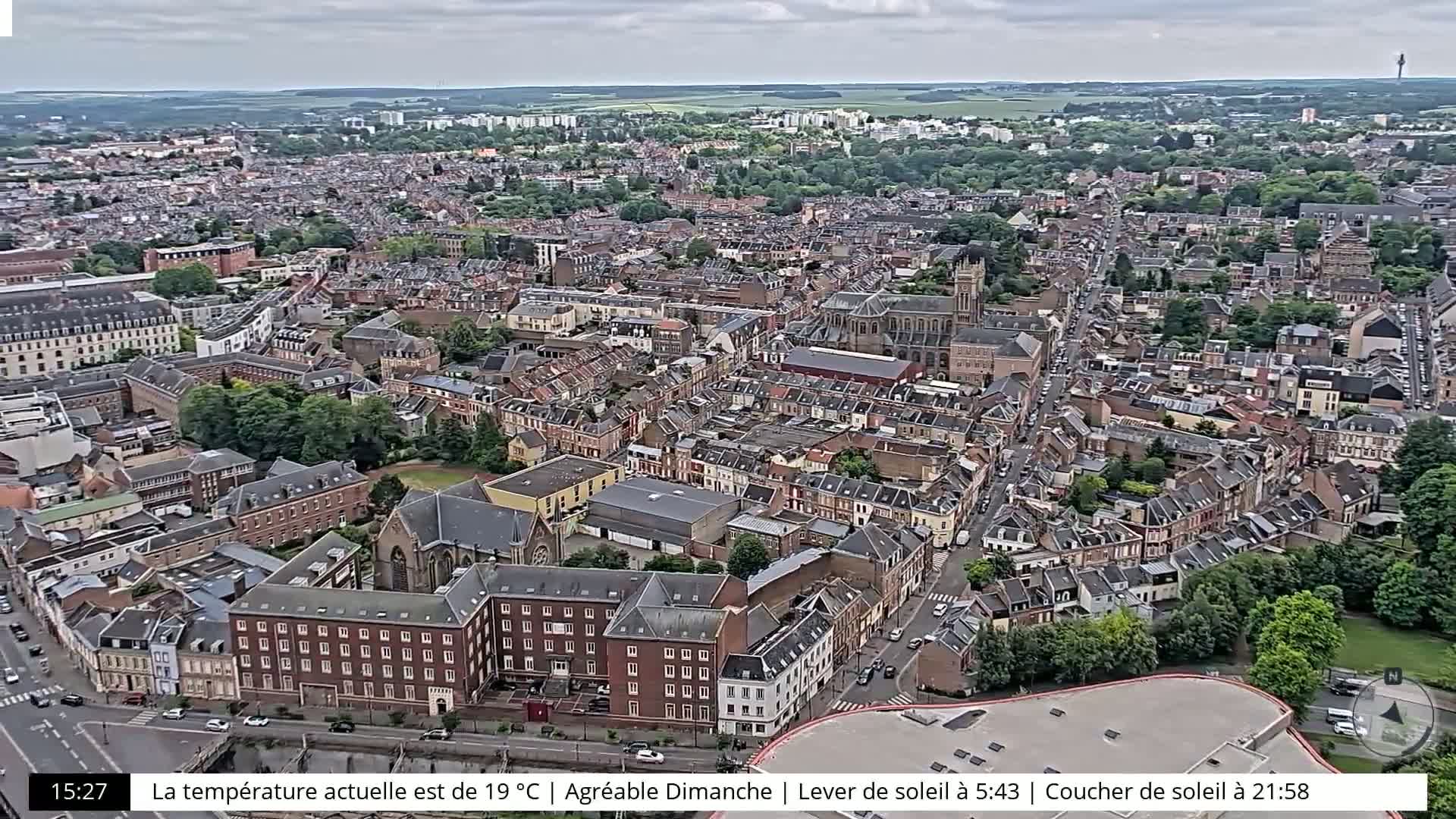 An aerial view of a city on an overcast day shows many brown-roofed buildings, green trees, and streets with cars.