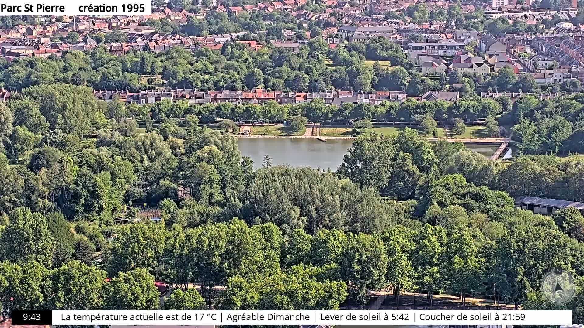 A high-angle, sunny view of a park with a pond, surrounded by lush green trees and residential buildings.