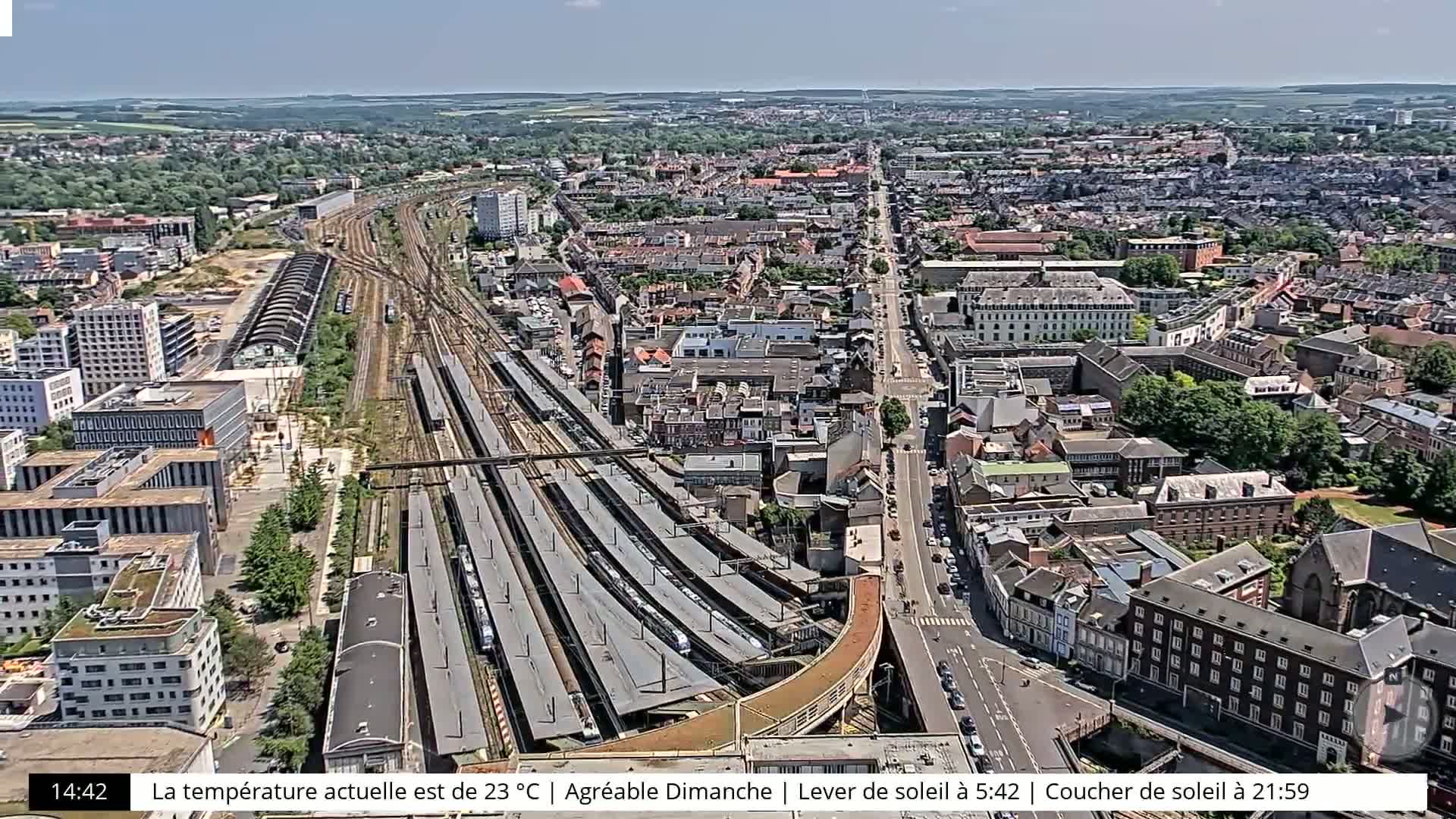 A high-angle, sunny view of a city's railway station and surrounding buildings and streets.