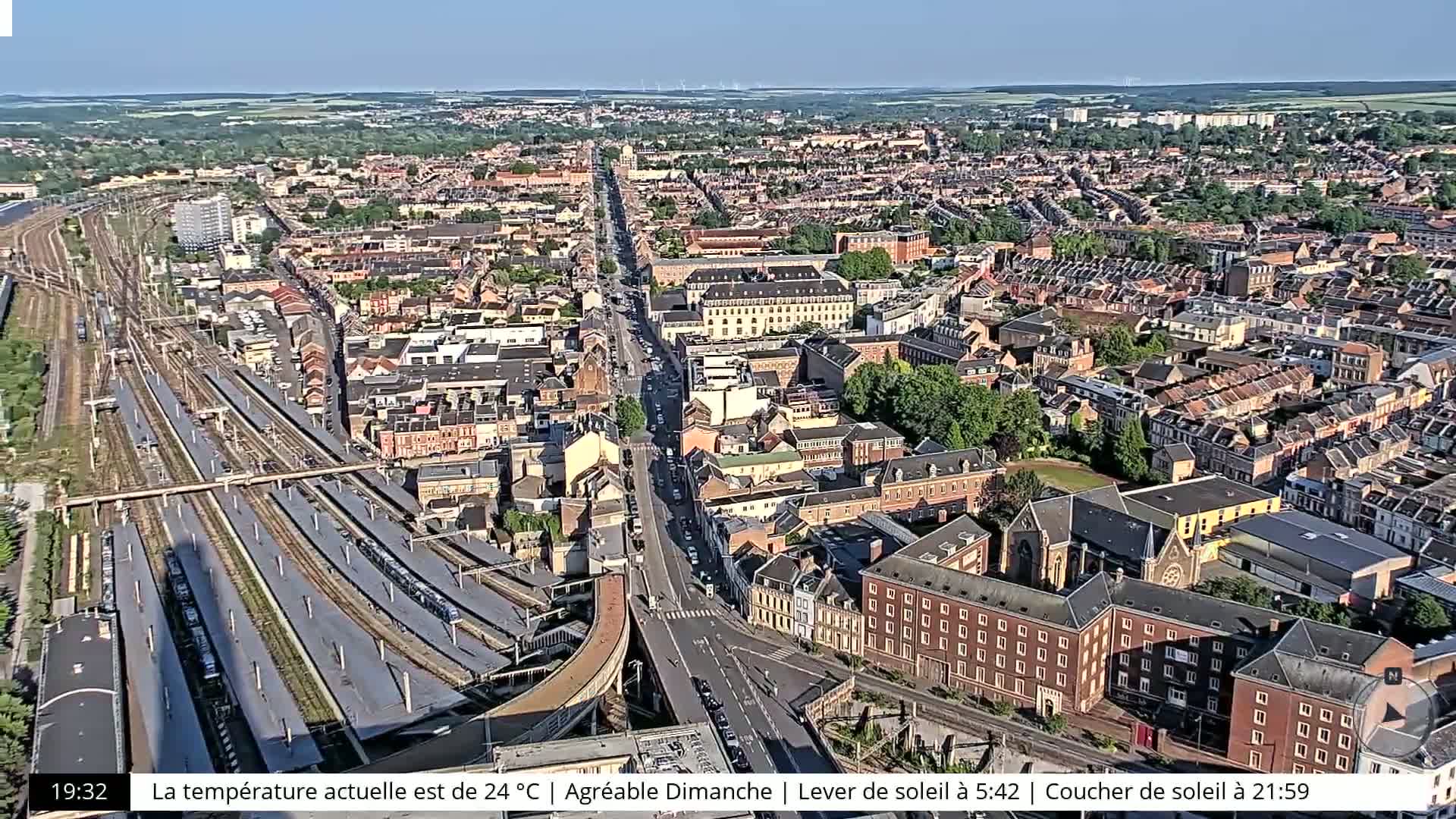 An aerial view of a city on a sunny day shows a large train station and numerous residential buildings.