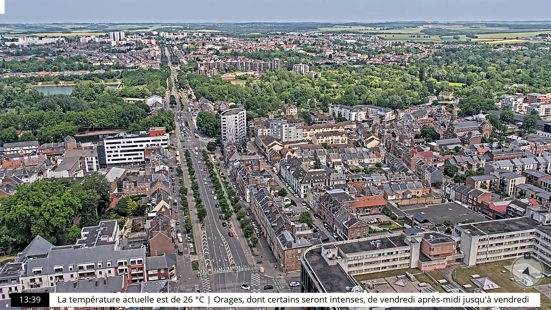 An aerial view shows a city with a wide road lined with trees, surrounded by residential buildings and green spaces under a clear sky.