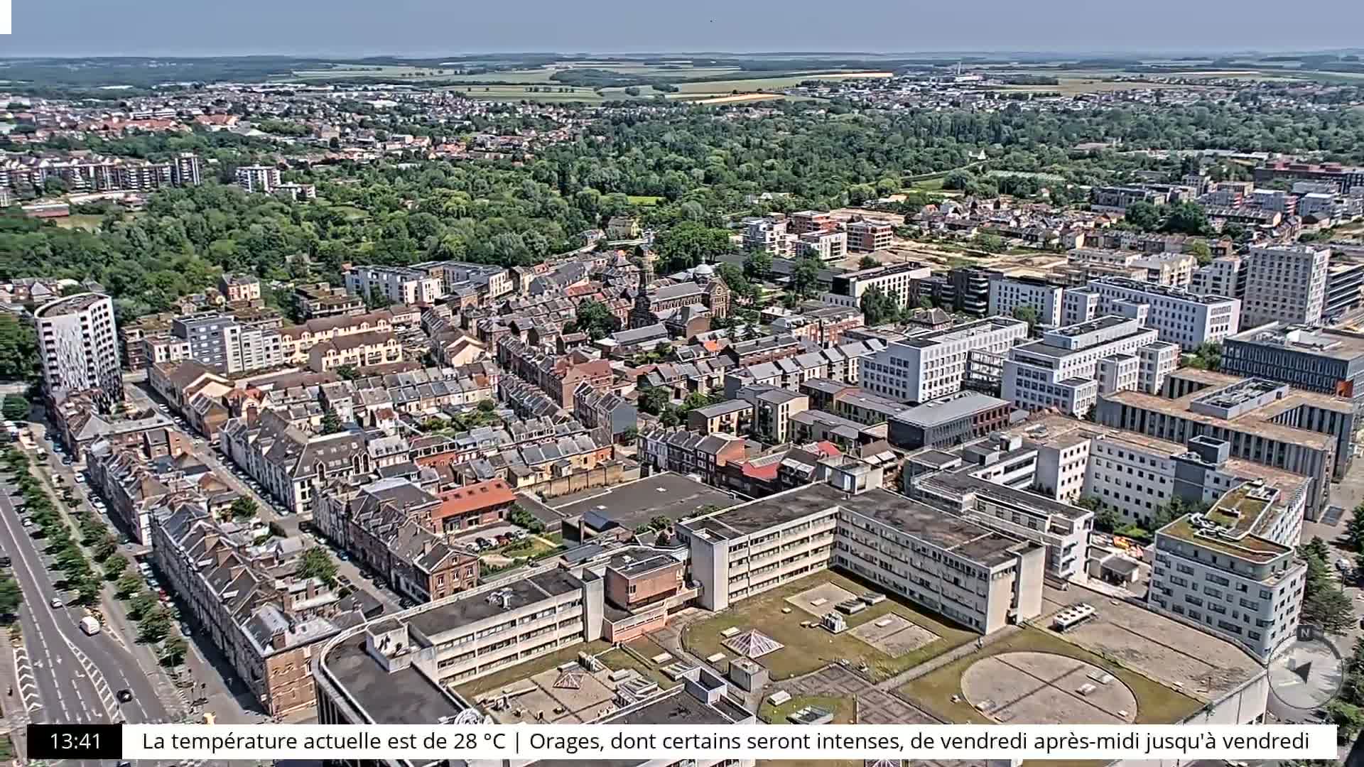 An aerial view of a city on a partly sunny day shows numerous buildings, streets, and green spaces.