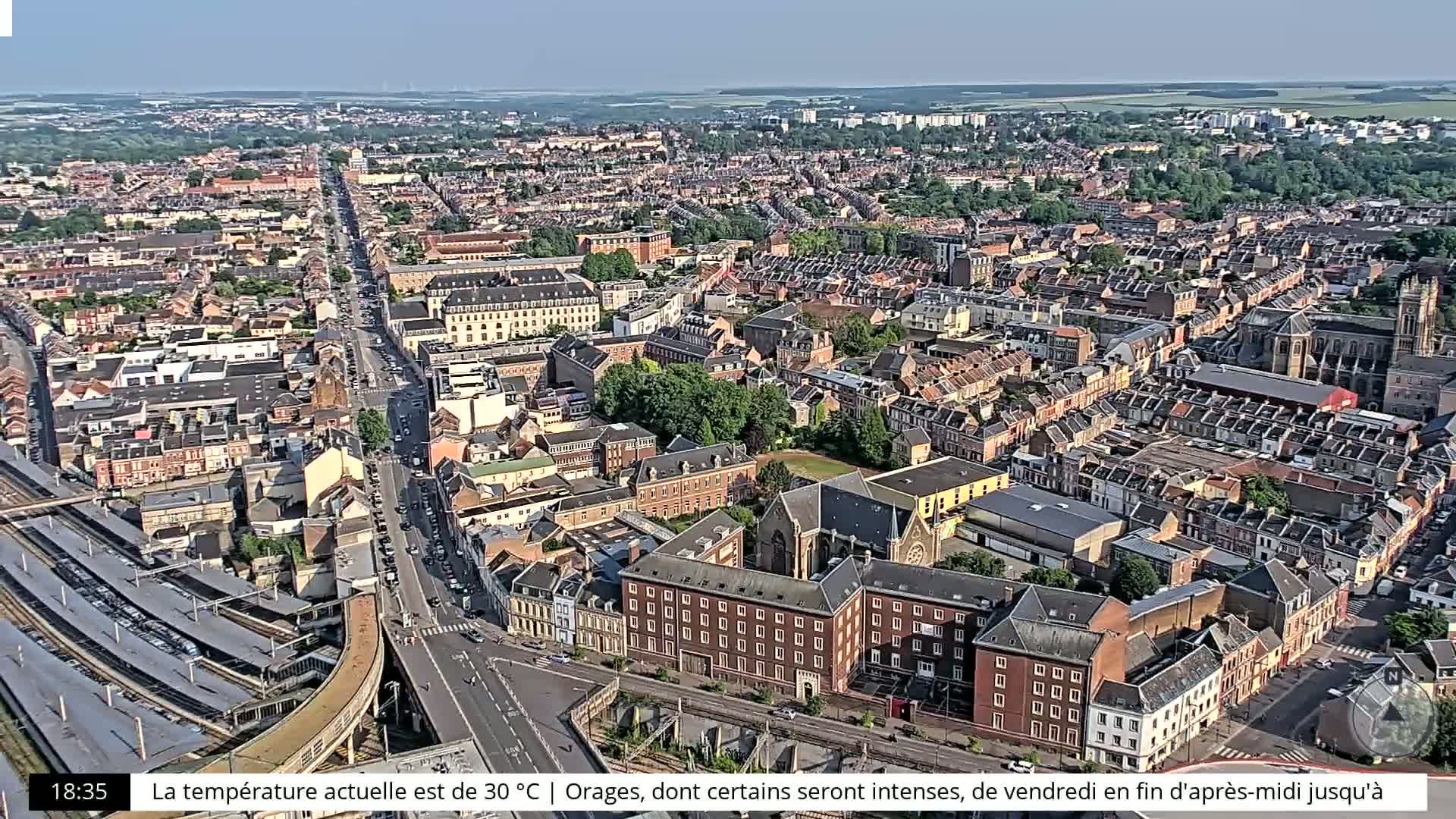 A sprawling cityscape with primarily brick buildings, a train station, and a major road running through it, under a clear sky.