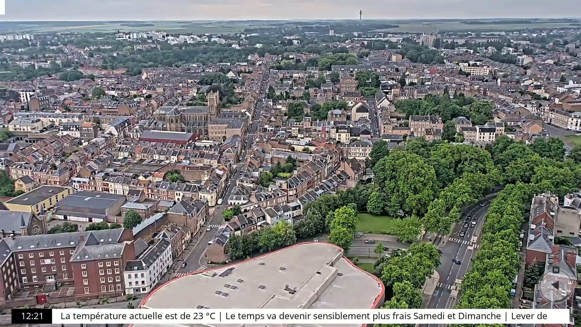 An aerial view shows a densely populated city under an overcast sky, with a large park and a large flat-roofed building visible.