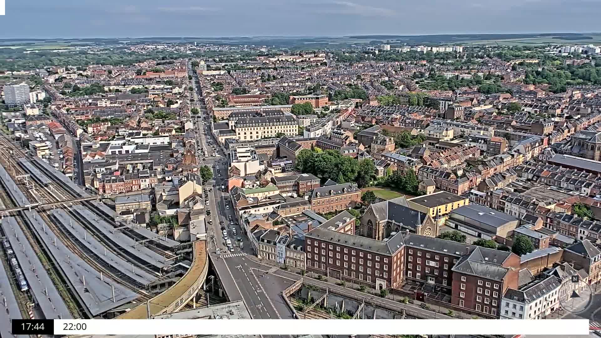 An aerial view of a densely populated city on a partly cloudy day, showing a train station and many residential buildings.