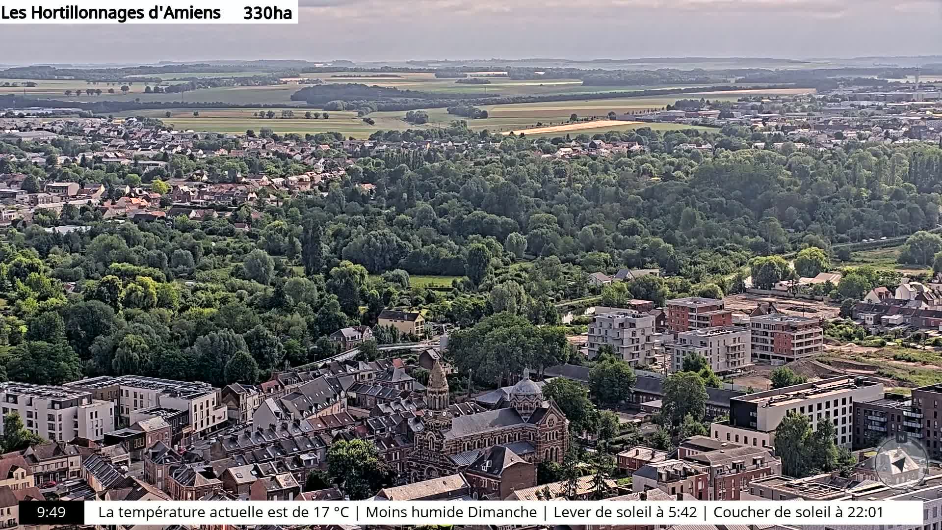 An aerial view shows a town with a mix of residential buildings, a large church, and extensive green areas, under a partly cloudy sky.