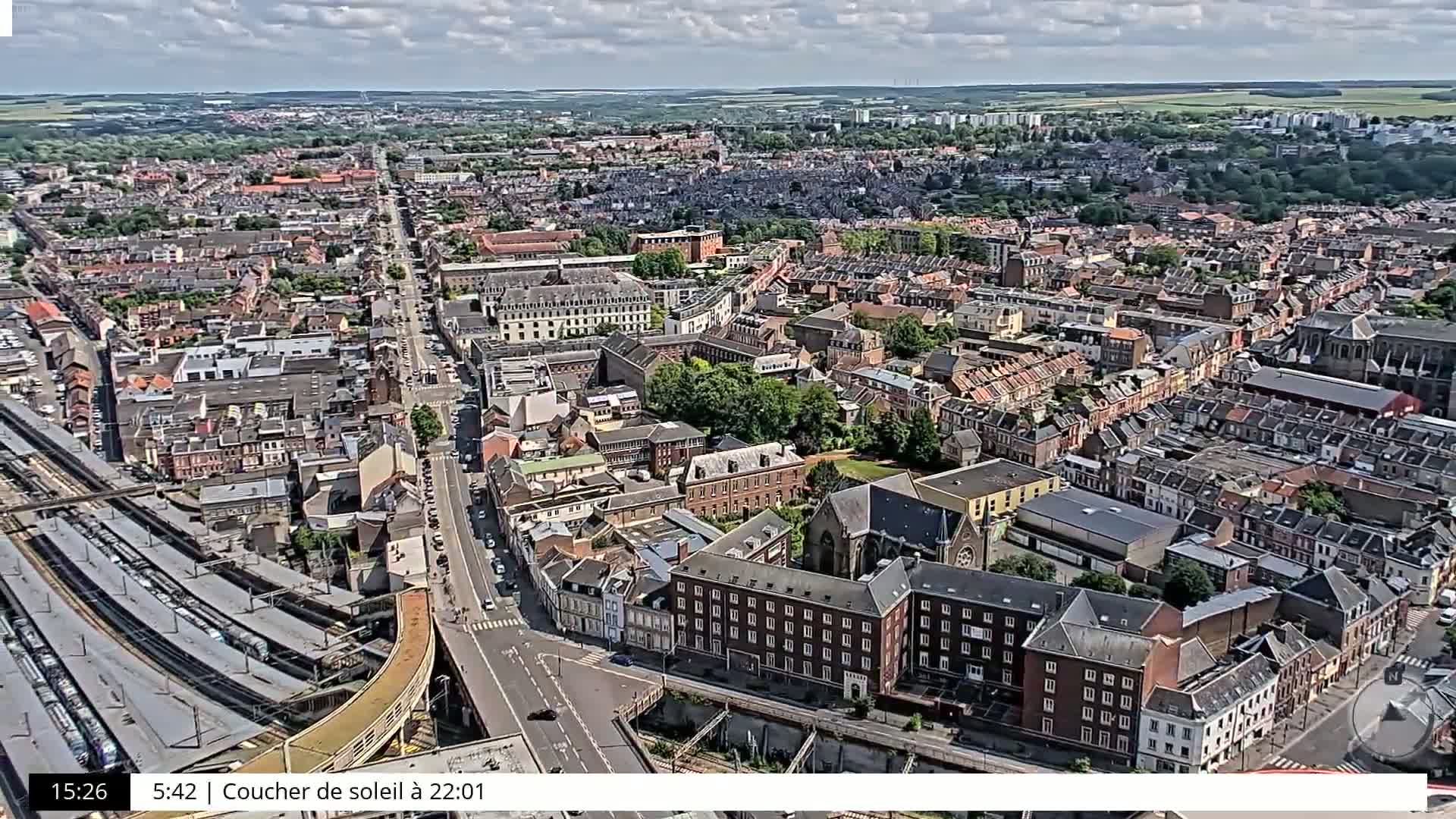 An aerial view of a city on a partly cloudy day shows a dense network of streets, buildings, and a train station.