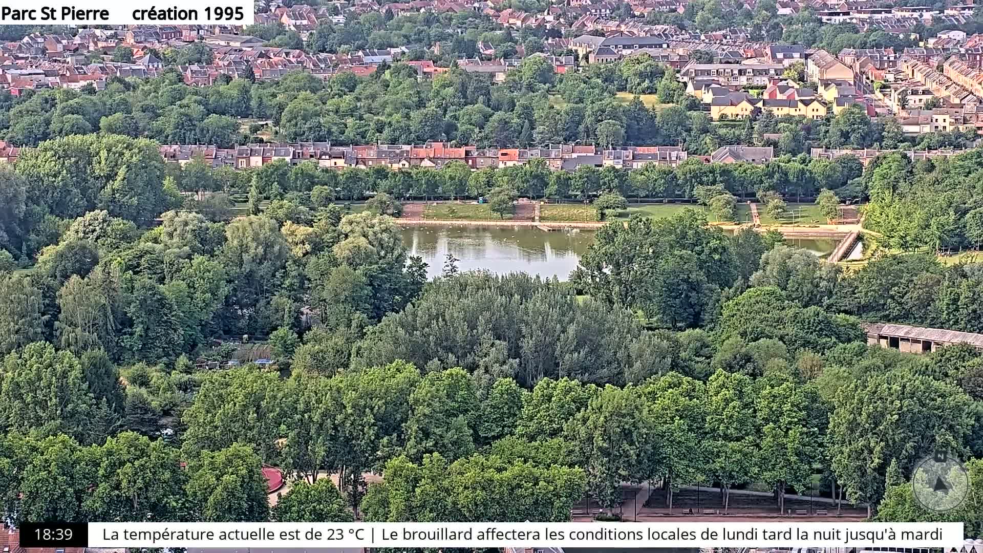 An aerial view of a park with a pond, surrounded by lush green trees and residential buildings in the background.