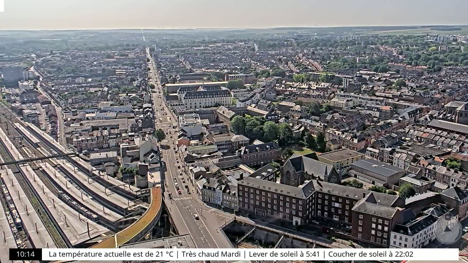 A high-angle, long shot of a city on a sunny day shows a wide street with train tracks to the left, and many buildings surrounding the street.