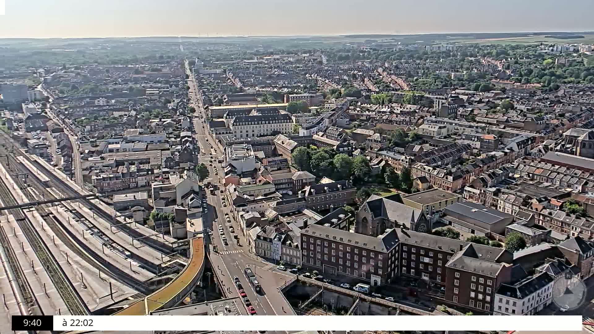 A high-angle, long shot on a sunny day shows a sprawling city with many closely packed buildings, a large road, and train tracks.