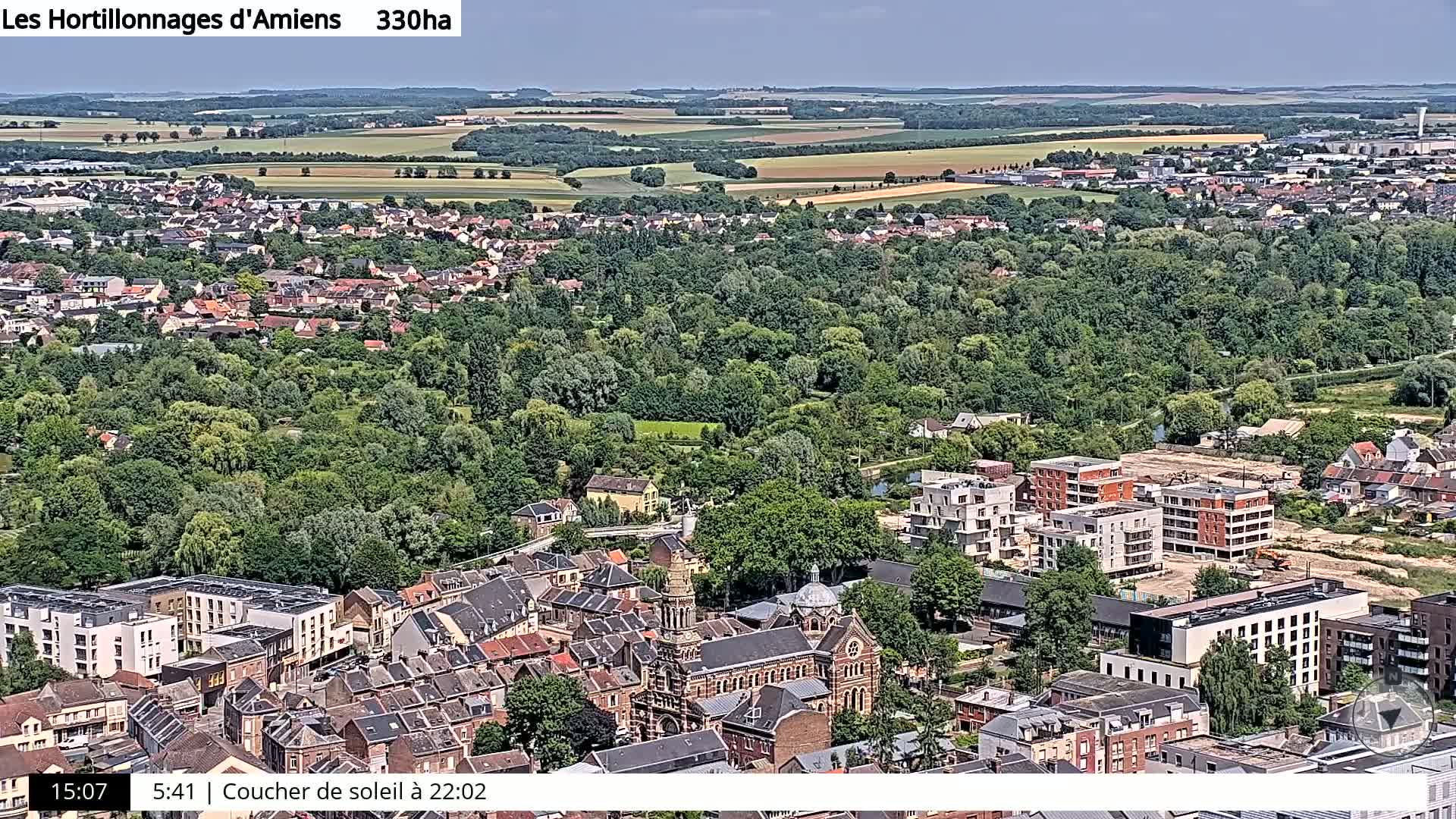 An aerial view shows a town with a mix of residential buildings, a large green area, and farmland in the distance under a mostly clear sky.