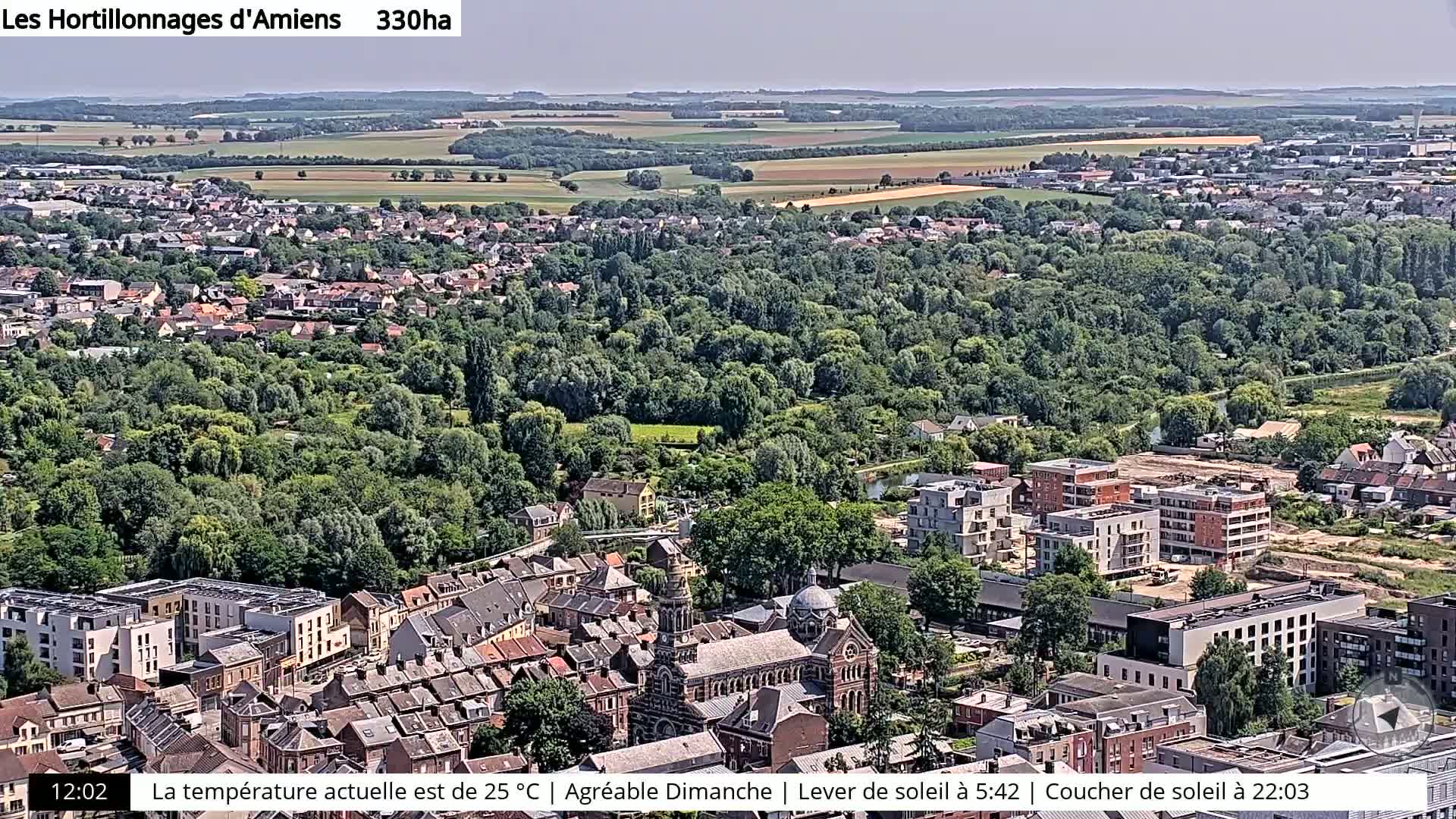 An aerial view shows a town with a mix of older and newer buildings, surrounded by lush green trees and farmland under a clear sky.