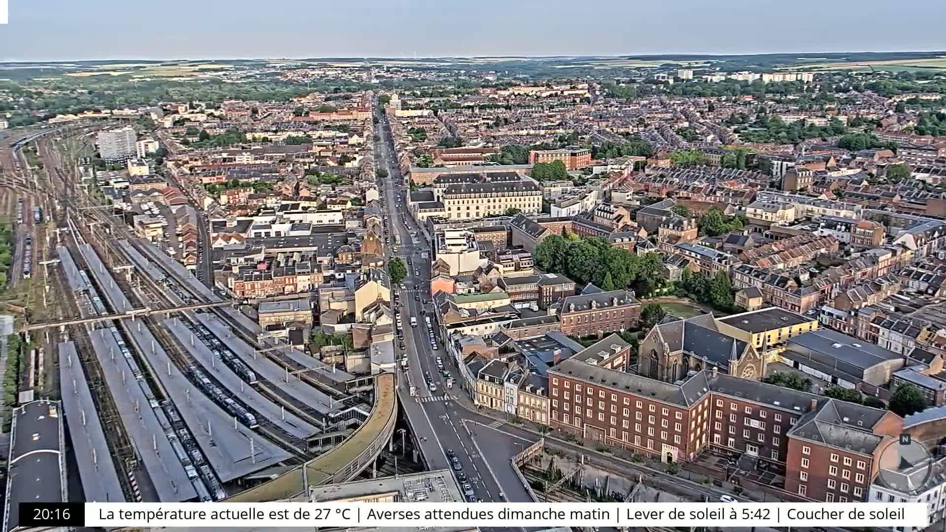 An aerial view shows a sprawling city with numerous buildings, train tracks filled with multiple trains, and a long road cutting through the center, all under a clear sky.