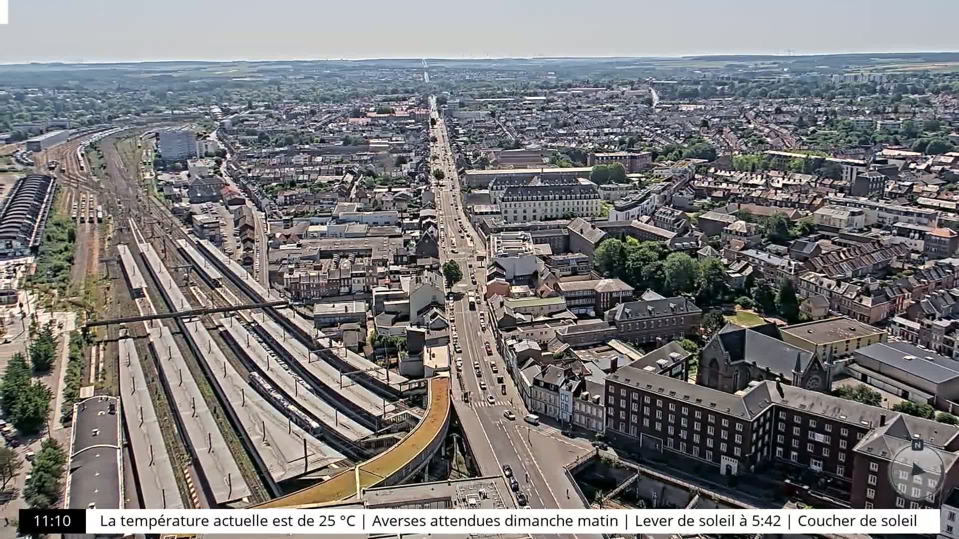 A high-angle, long shot shows a city with a train station and a main road running through it on a sunny day.