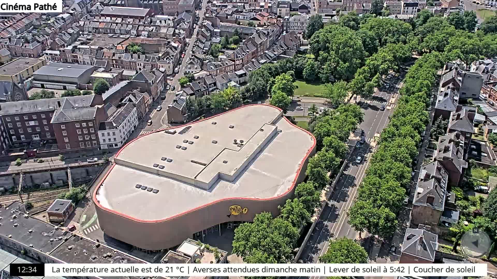An aerial view shows a large, modern building with a curved roof situated amongst smaller buildings and a tree-lined street on a sunny day.