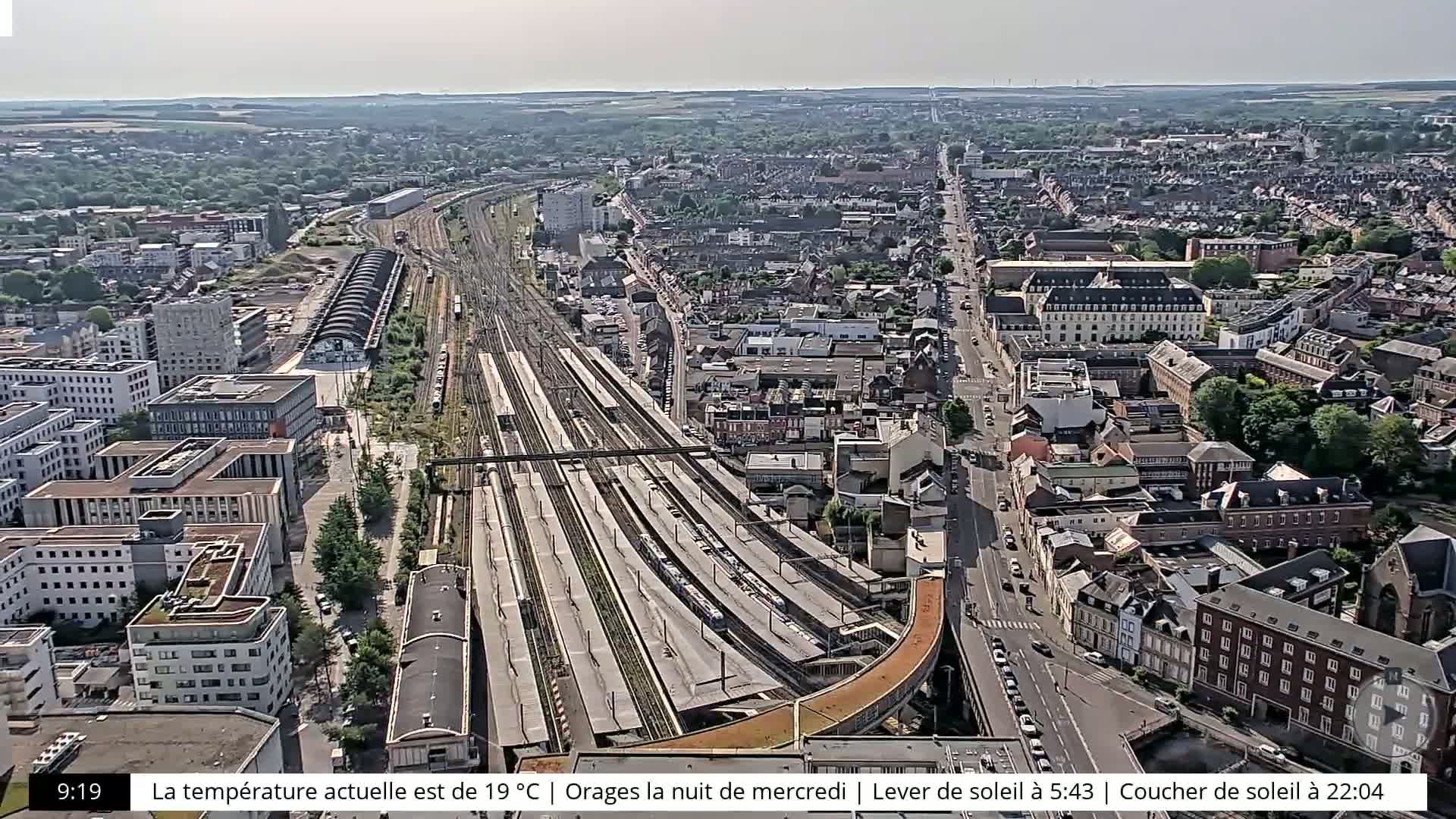 A high-angle, sunny view of a city with a large train station and numerous buildings, extending to a horizon of greenery.