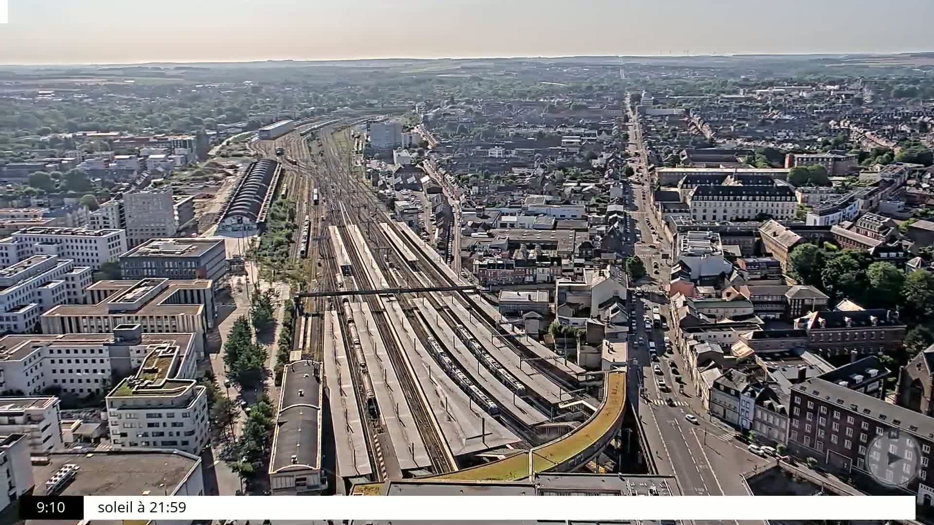 A high-angle, sunny view of a city with a large train station and numerous buildings, roads, and trees.