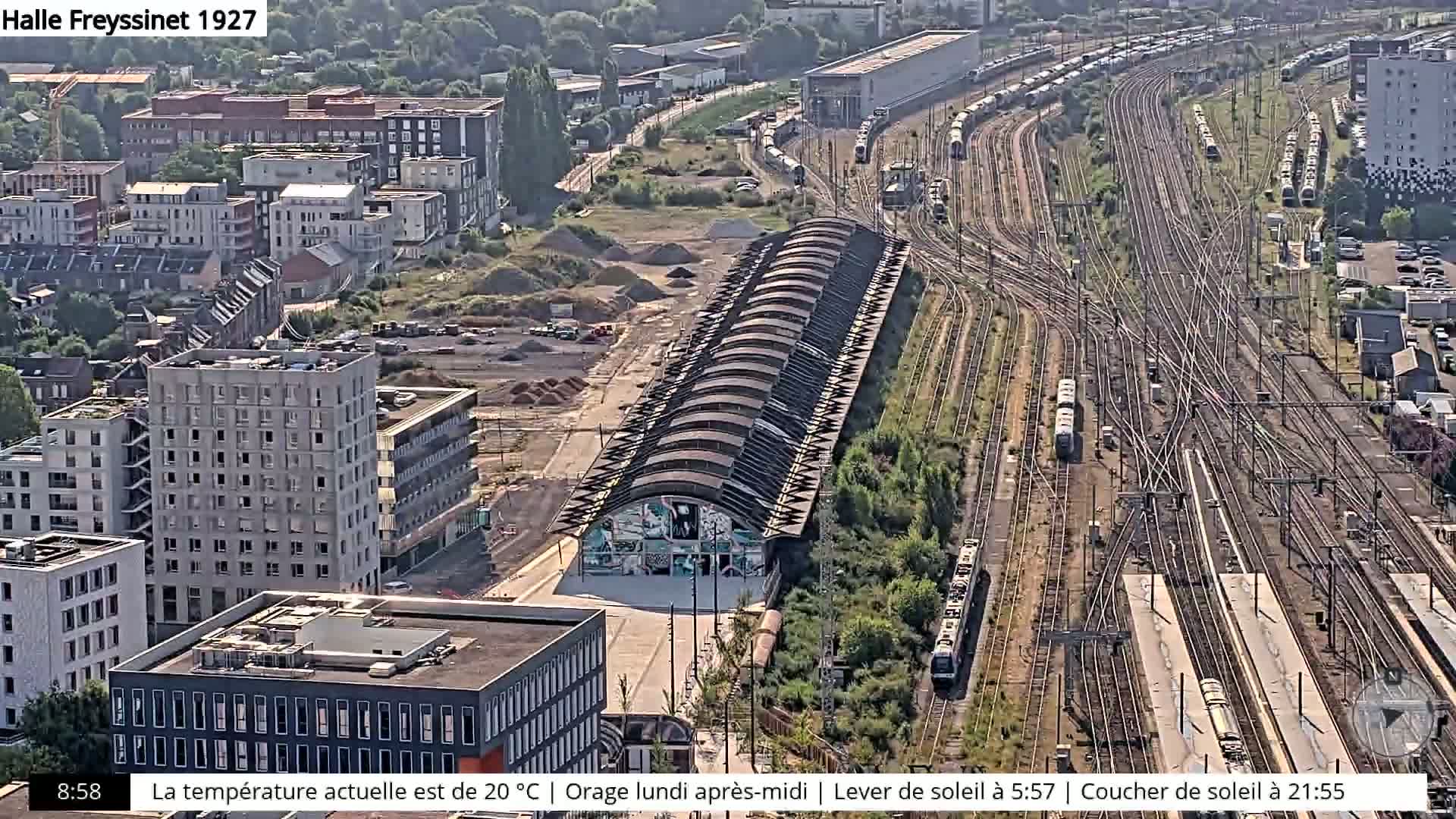 An aerial view shows a large train yard with numerous tracks and parked trains, alongside a row of arched buildings and a developing urban area with modern apartment buildings, under a partly sunny sky.