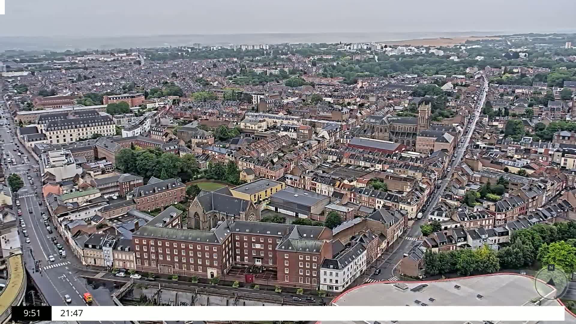 A sprawling city, under an overcast sky, is viewed from above, showing a dense network of streets and buildings, with a large church visible in the center.