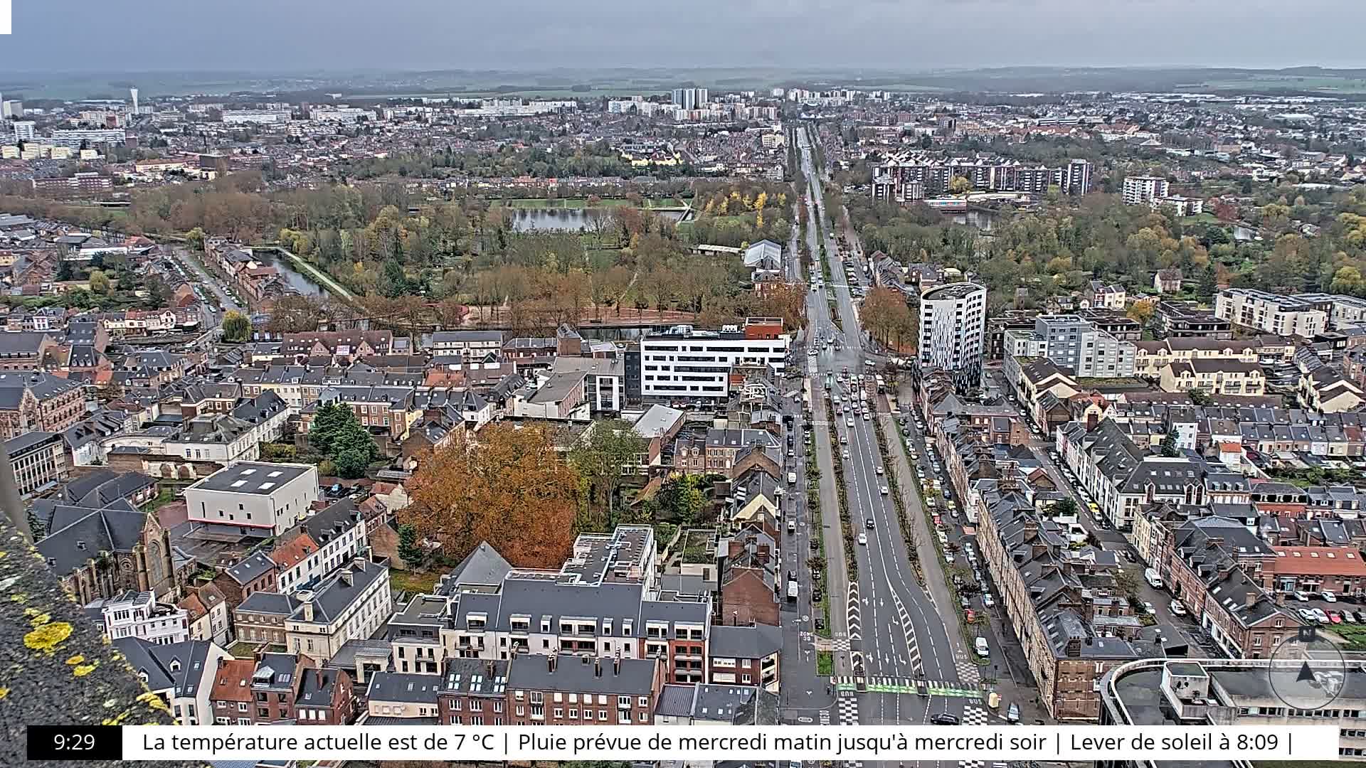 An expansive aerial view captures a sprawling urban landscape under a uniformly overcast sky, featuring a long, tree-lined avenue bustling with cars, a mix of traditional and modern buildings, and a central park with a pond amidst trees showing autumnal foliage.