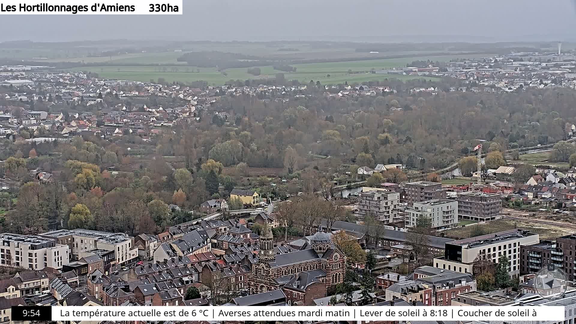 A panoramic view captures a town with a mix of residential and modern buildings, a prominent church, and a large expanse of mostly leafless trees and fields in the distance, all under an overcast sky.