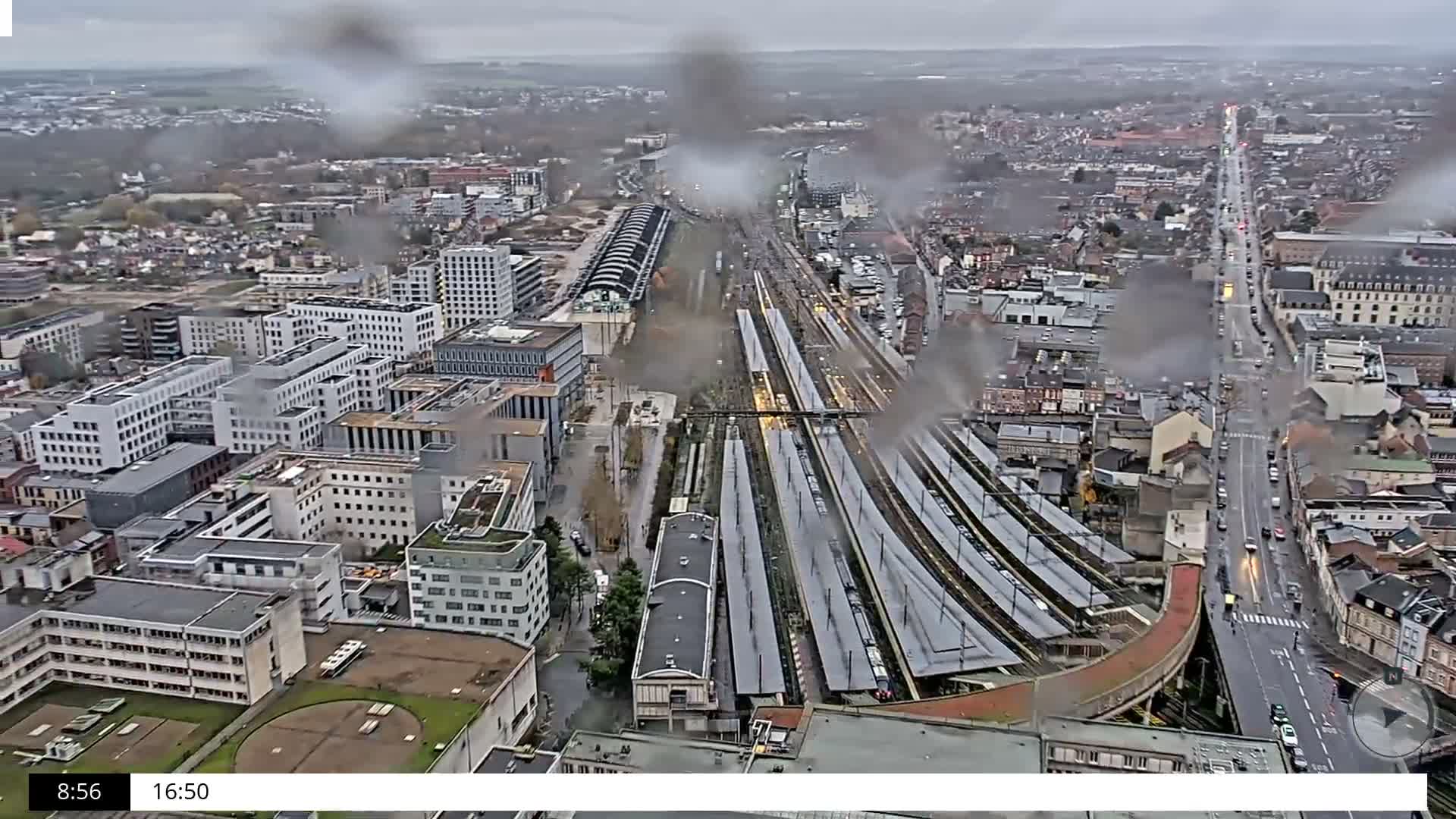 An overcast and rainy aerial view captures a large train station with multiple tracks and platforms at the center of an urban landscape, flanked by diverse city buildings and busy streets.