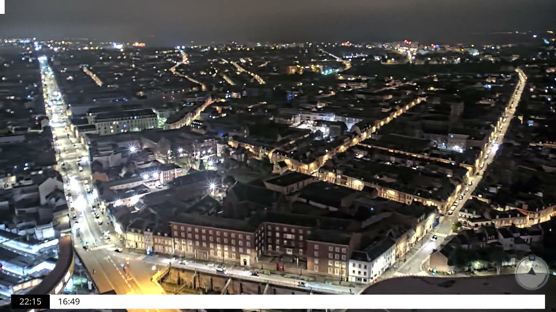 An overcast and rainy aerial view captures a large train station with multiple tracks and platforms at the center of an urban landscape, flanked by diverse city buildings and busy streets.