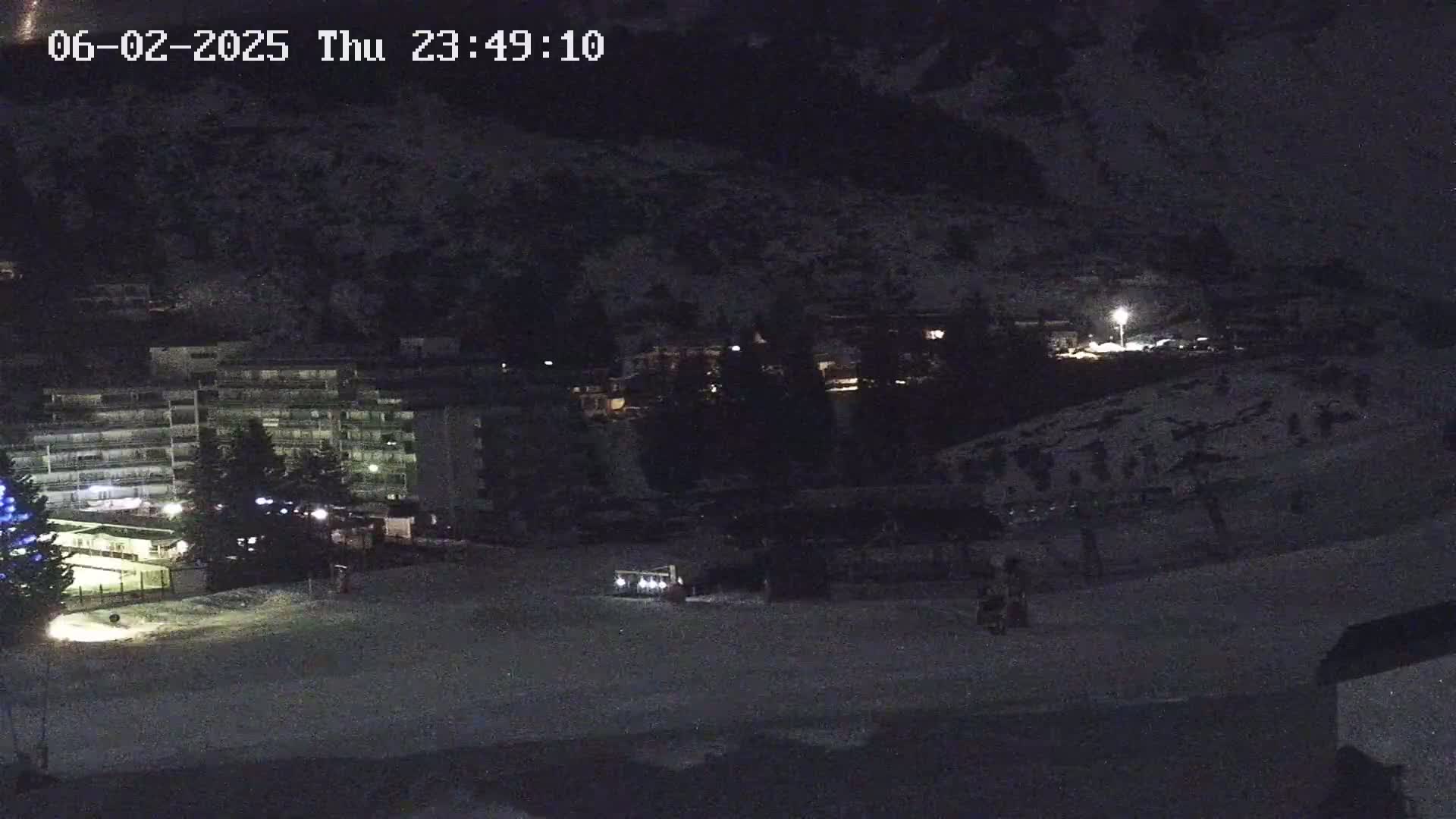 A nighttime, snow-covered scene shows a multi-story building, some smaller structures, and a dark hillside under a clear, dark sky.