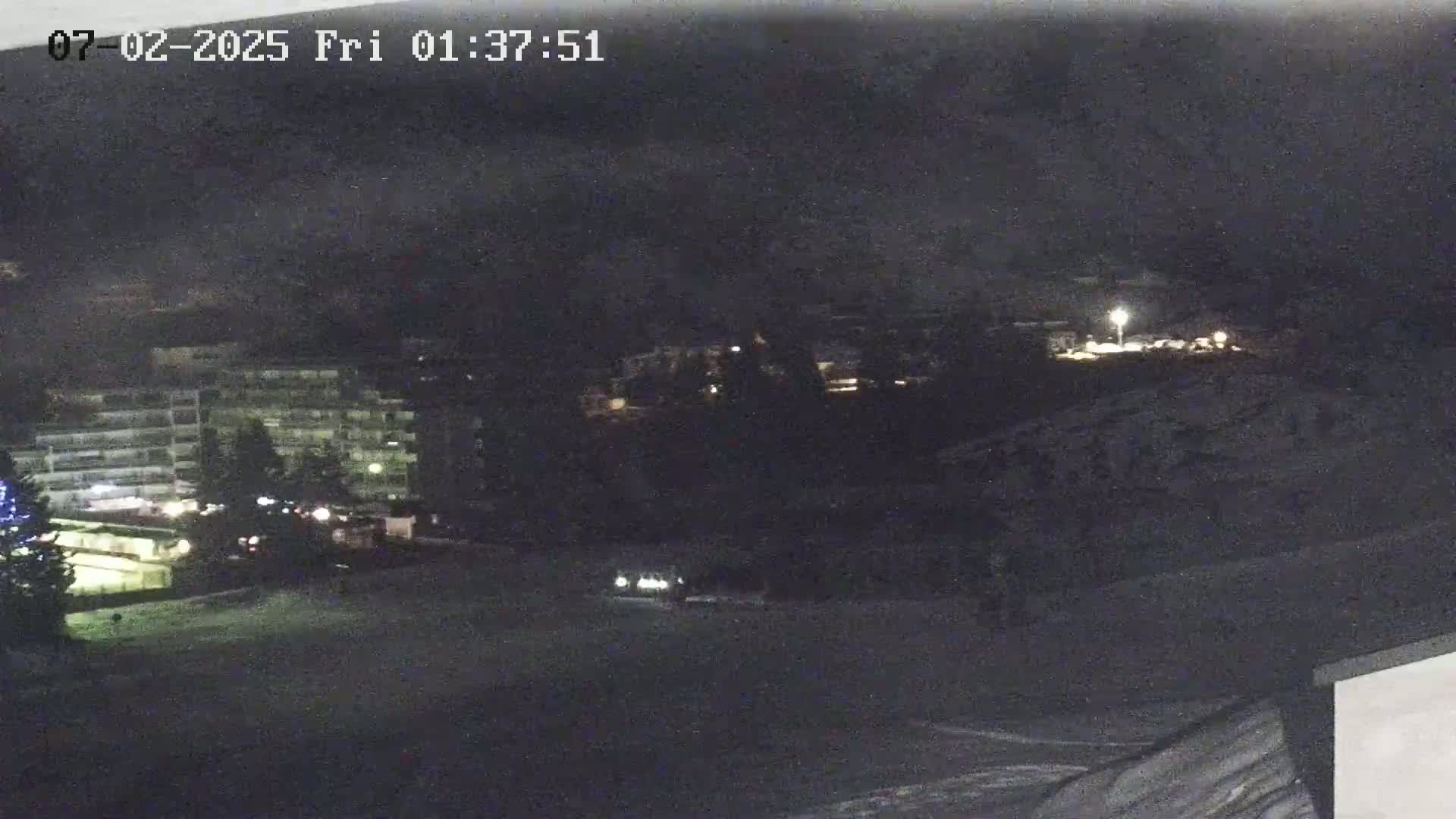 A nighttime view of a snow-covered landscape shows a multi-story building, some vehicles, and sparsely lit areas.
