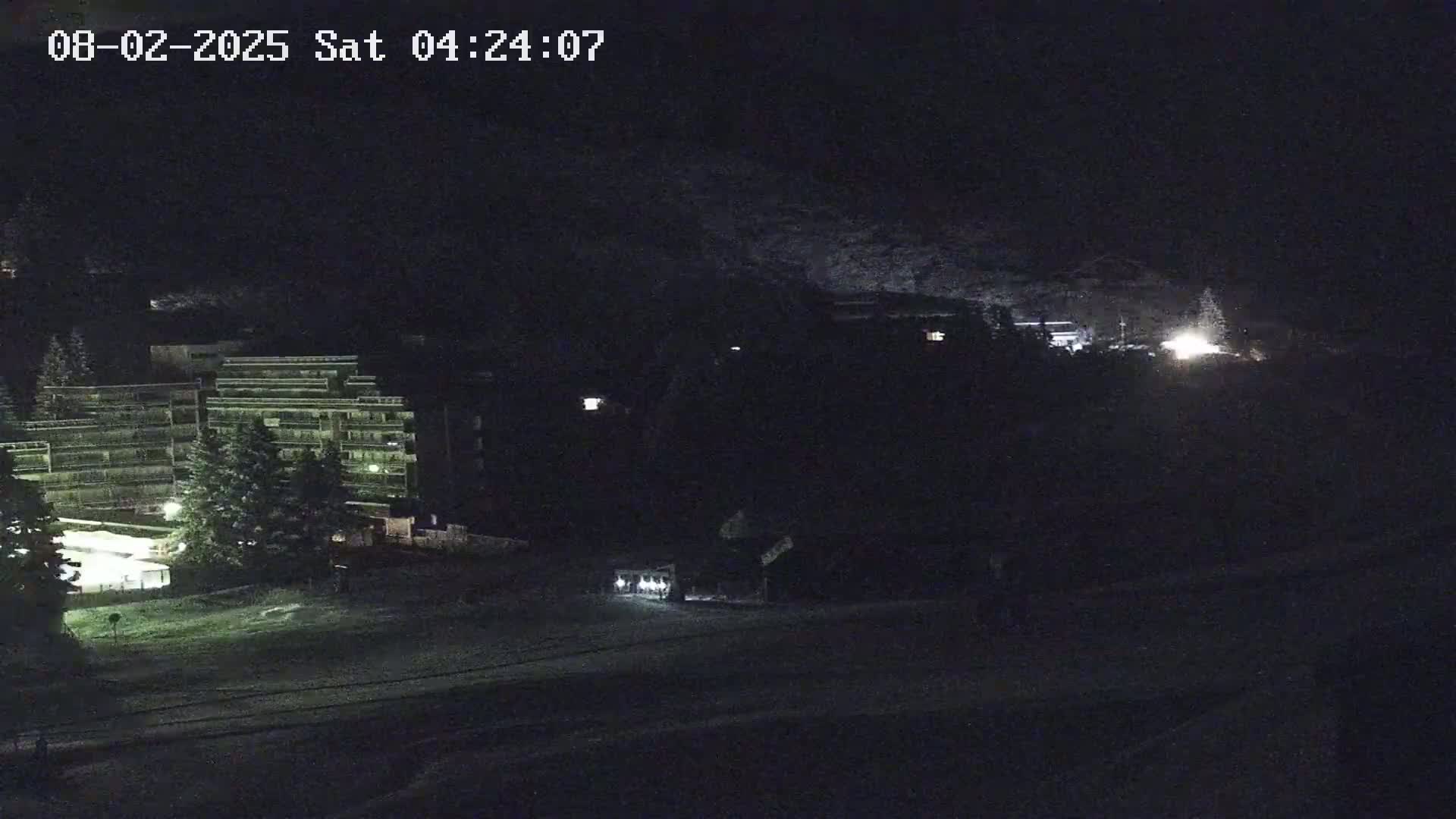 A nighttime, snow-covered scene shows a multi-story building and other structures illuminated by artificial light against a dark, mountainous background.