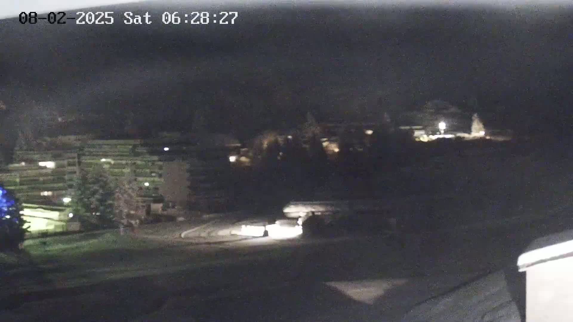 A nighttime view of a parking lot with several cars and buildings, some with lights on, in the background.
