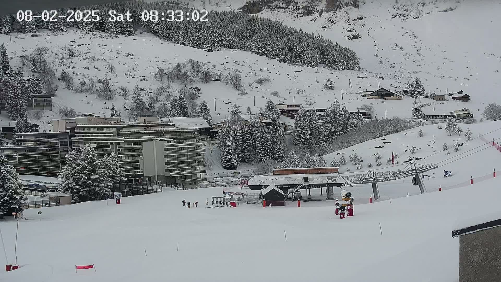A snow-covered ski resort with buildings, a ski lift, and several people skiing on a snowy slope under a cloudy sky.
