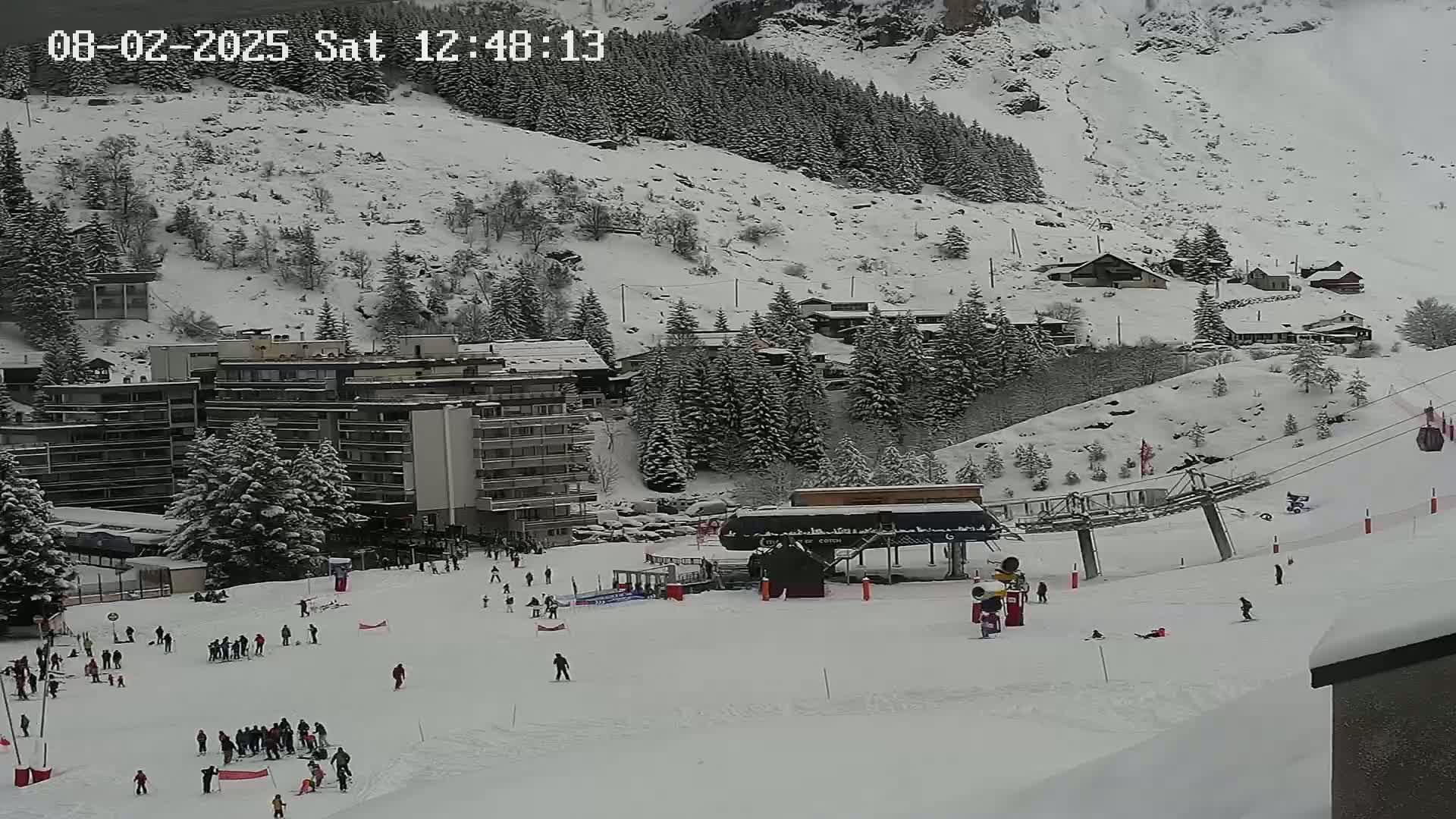 A snow-covered ski slope with skiers and buildings in the background under an overcast sky.