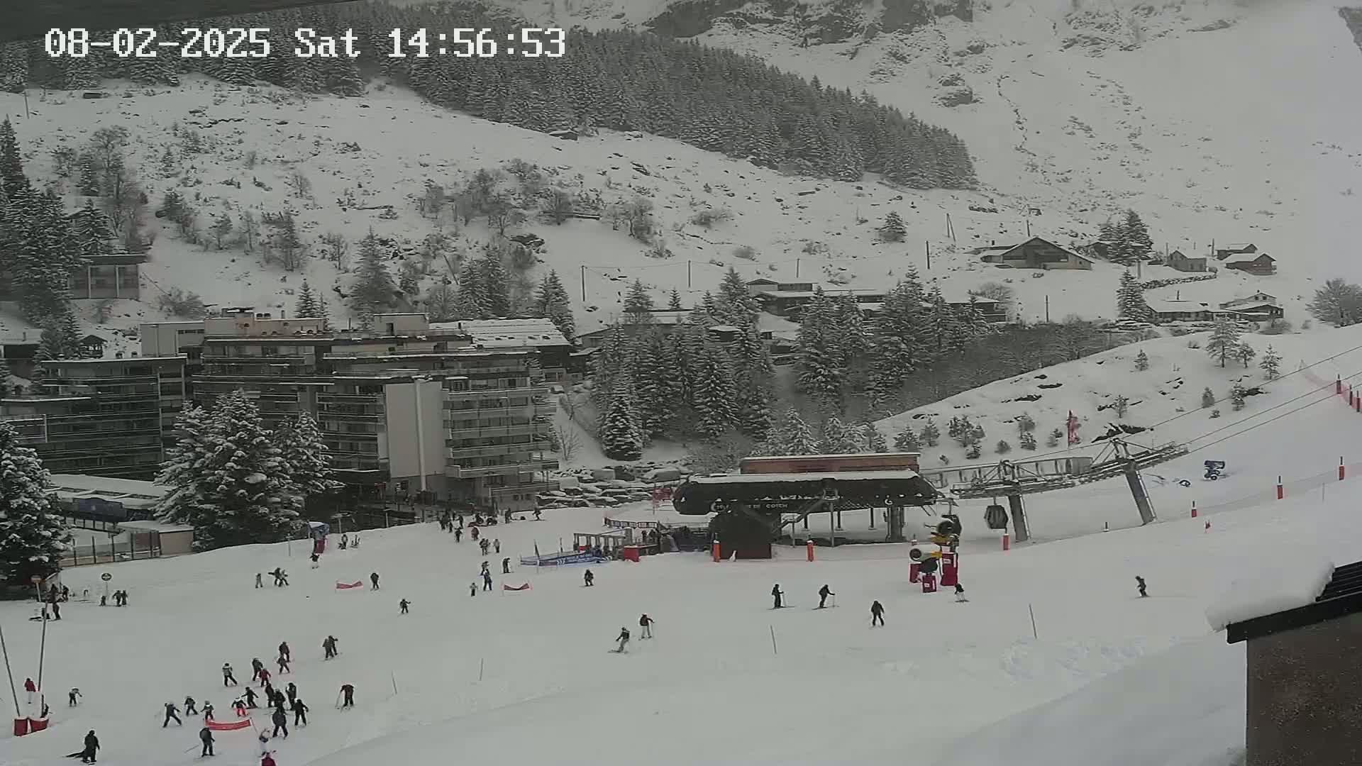 A snowy ski slope with a chairlift and several skiers and snowboarders, situated below a snow-covered mountain and residential buildings in a light snow.