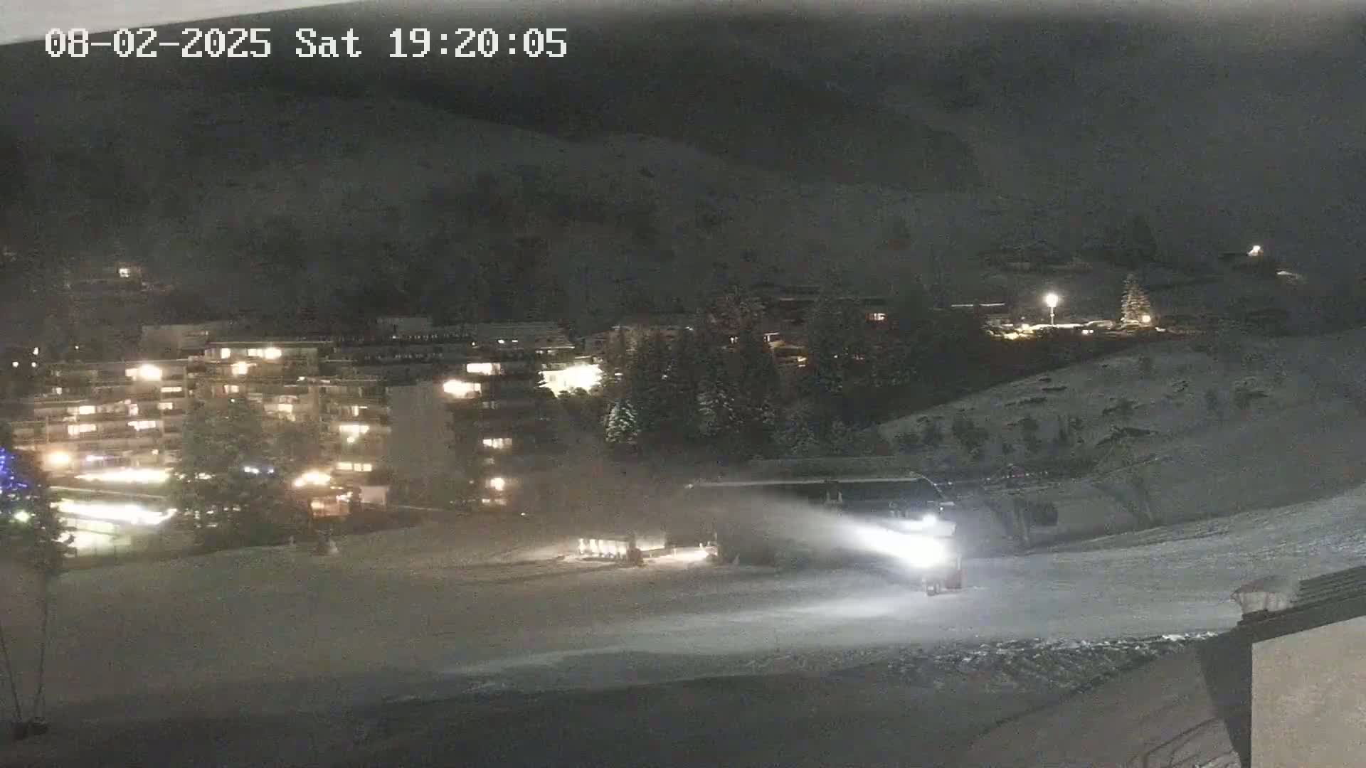 A snowcat illuminates a snow-covered slope at night near a brightly lit apartment building.