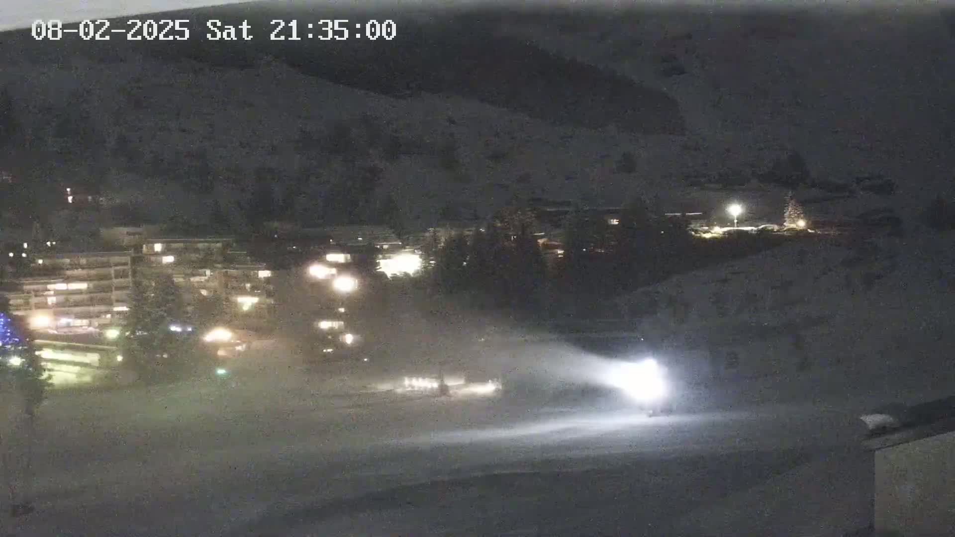 A snow groomer illuminates a snow-covered area at night near a building complex with lights on, under snowy conditions.