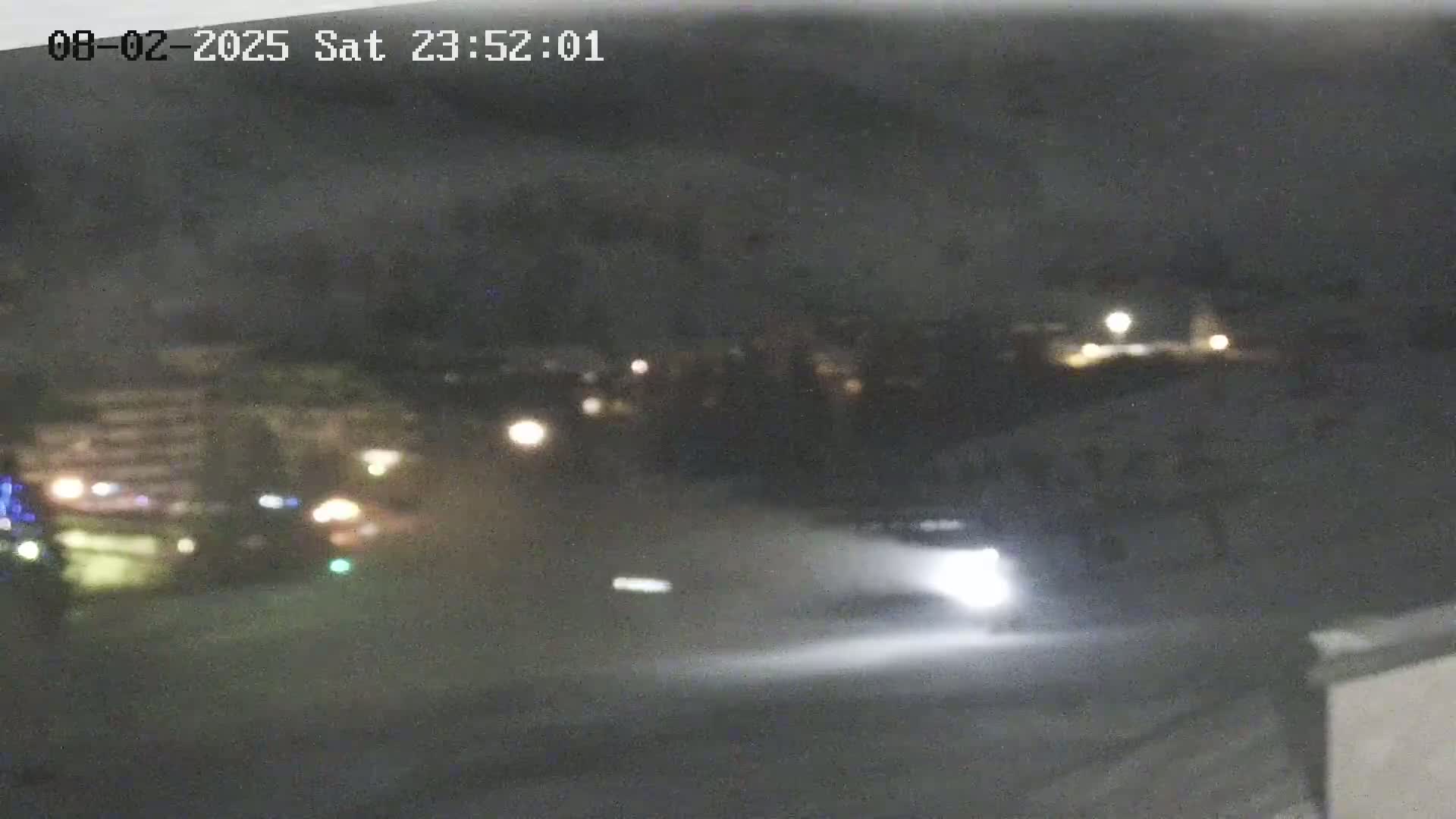 A nighttime, blurry view shows a vehicle's headlights illuminating a snowy or icy surface in front of a dimly lit town.
