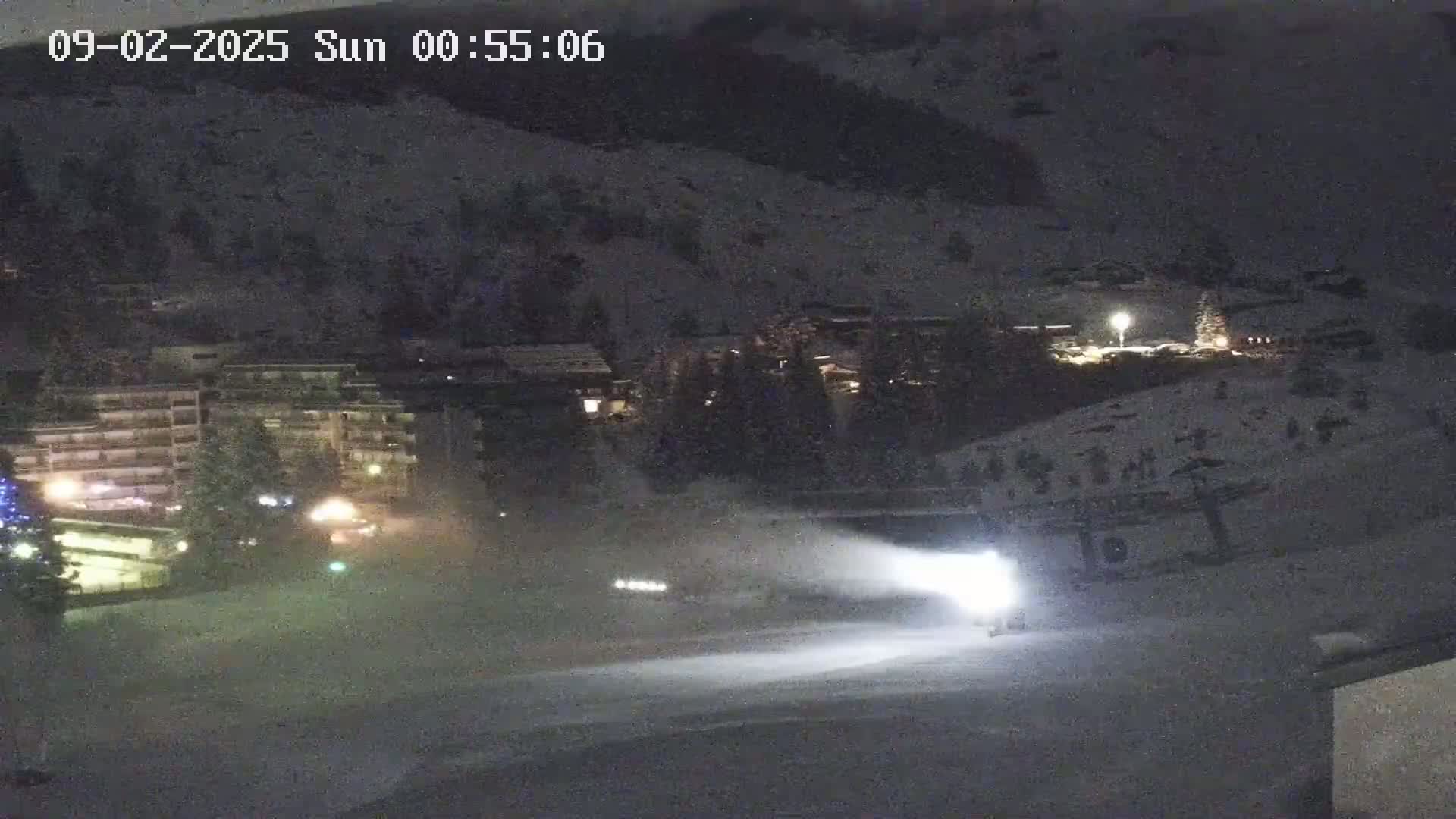 A snow groomer illuminates a snow-covered slope at night, with buildings and lights visible in the background.