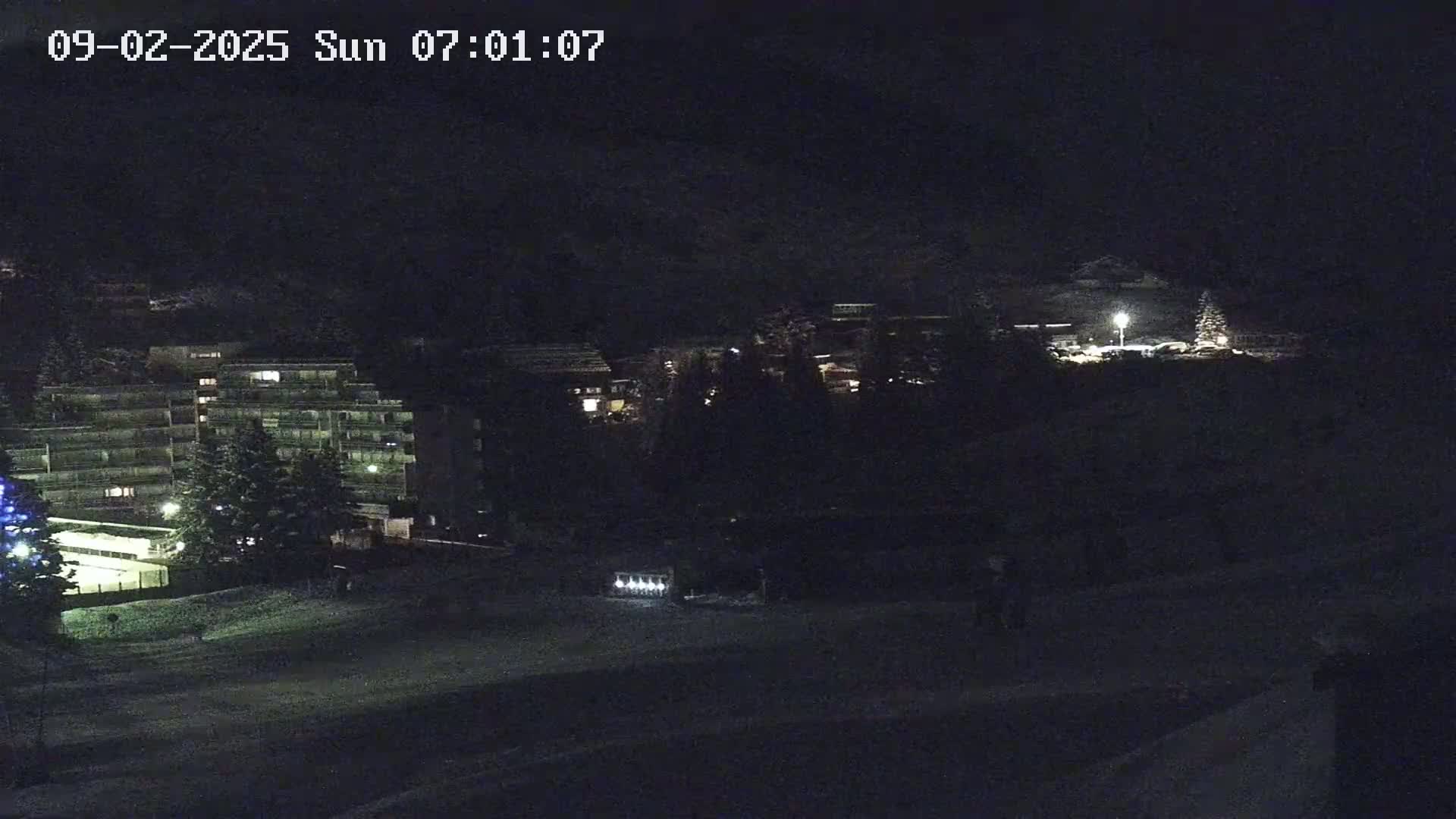 A nighttime view of snow-covered ground with buildings and trees illuminated by lights.