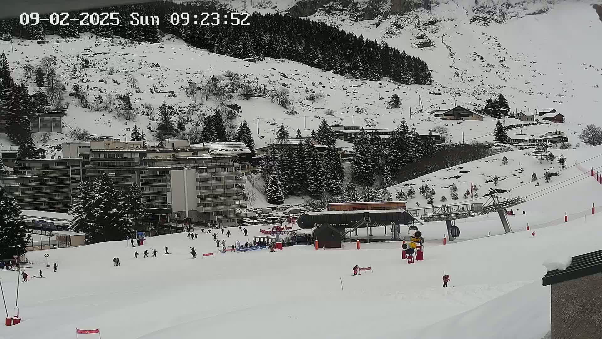 A snowy ski slope with several skiers, a ski lift, and buildings in the background, under overcast skies.