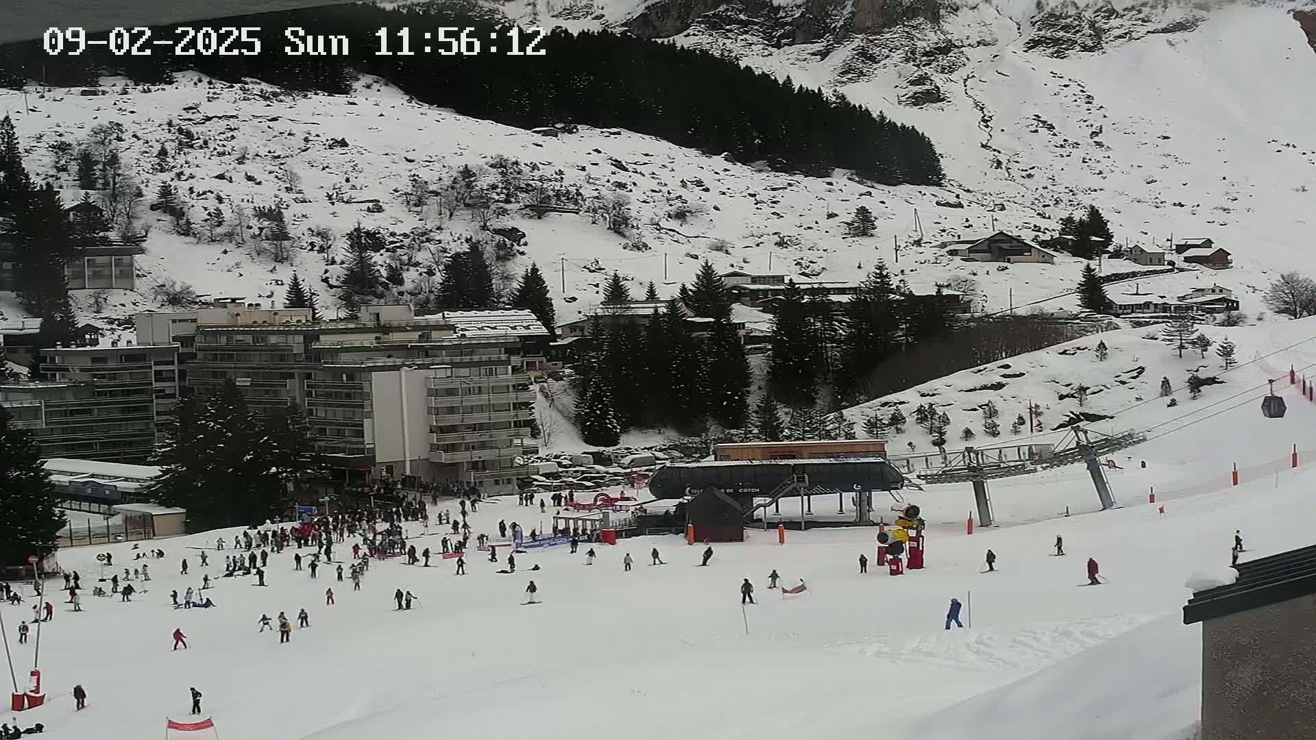 A snowy mountain slope with skiers and a ski lift, several buildings, and evergreen trees in the background under a cloudy sky.