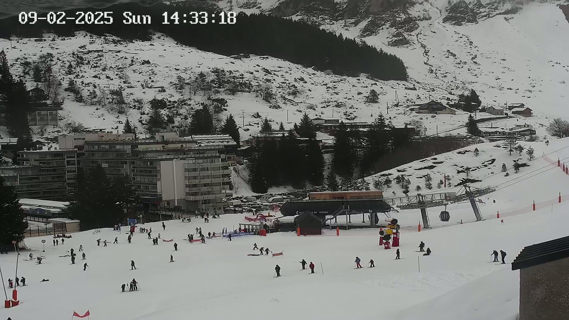 A snow-covered ski slope with skiers and a ski lift, situated in front of buildings and a snow-dusted mountainside.