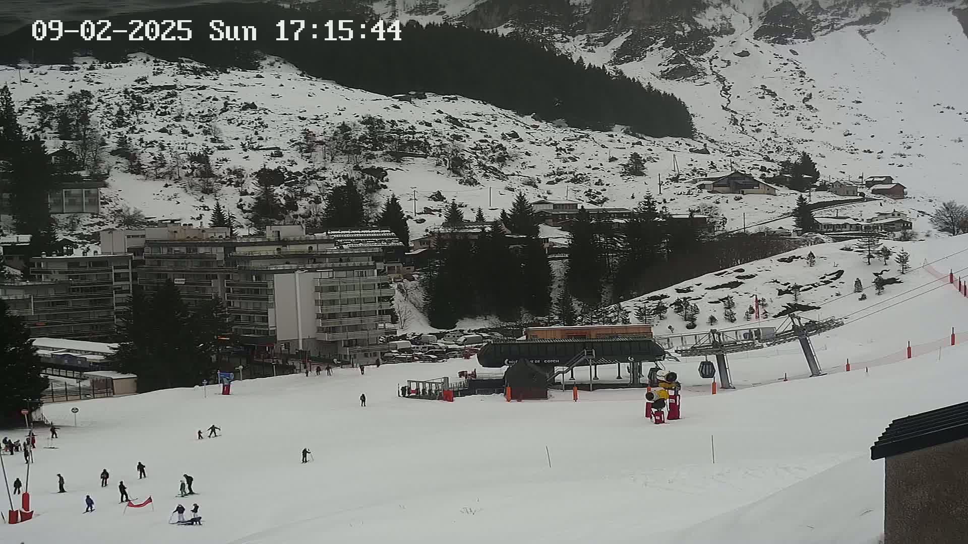 A snowy mountainside ski resort with several buildings, a ski lift, and skiers on a slope under an overcast sky.