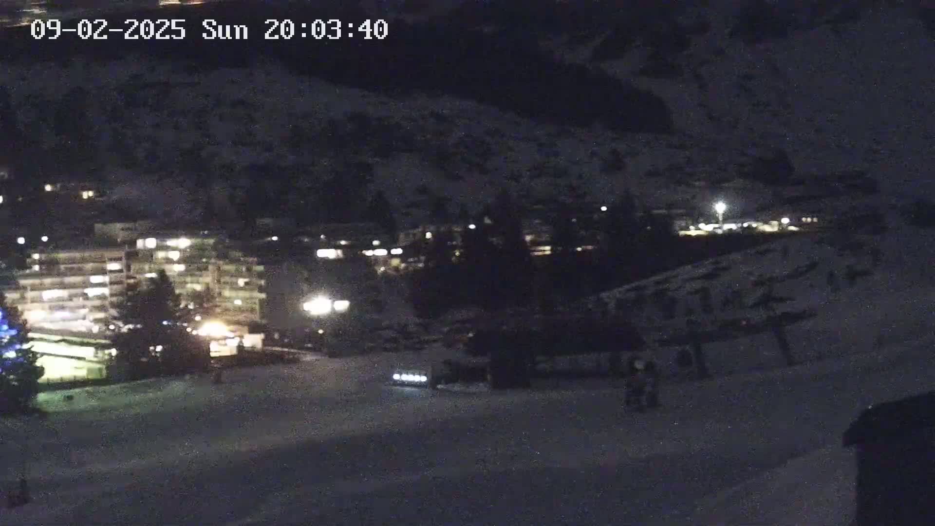 A nighttime, snow-covered scene shows a few buildings with illuminated windows, a road with a vehicle's headlights visible, and a few figures in the distance.