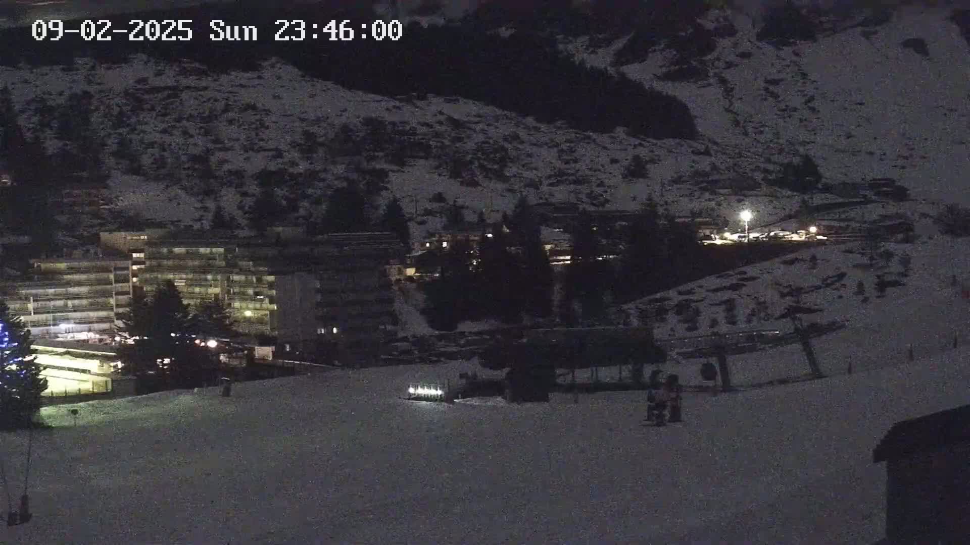 A snow-covered nighttime scene shows a ski lift, a multi-story building with lights on, and a few people near the bottom of a gently sloping hill.