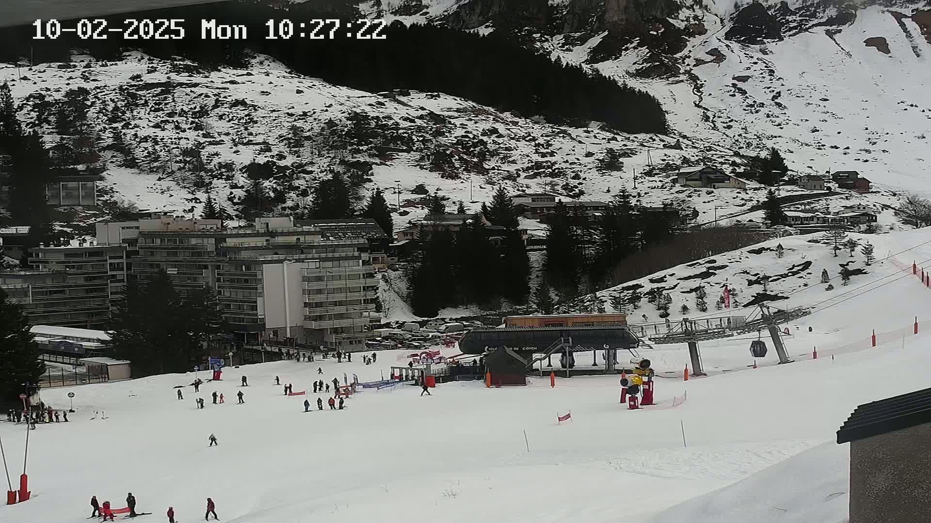 A snow-covered ski slope with several skiers and a chairlift, situated below a mountainside with buildings and trees.