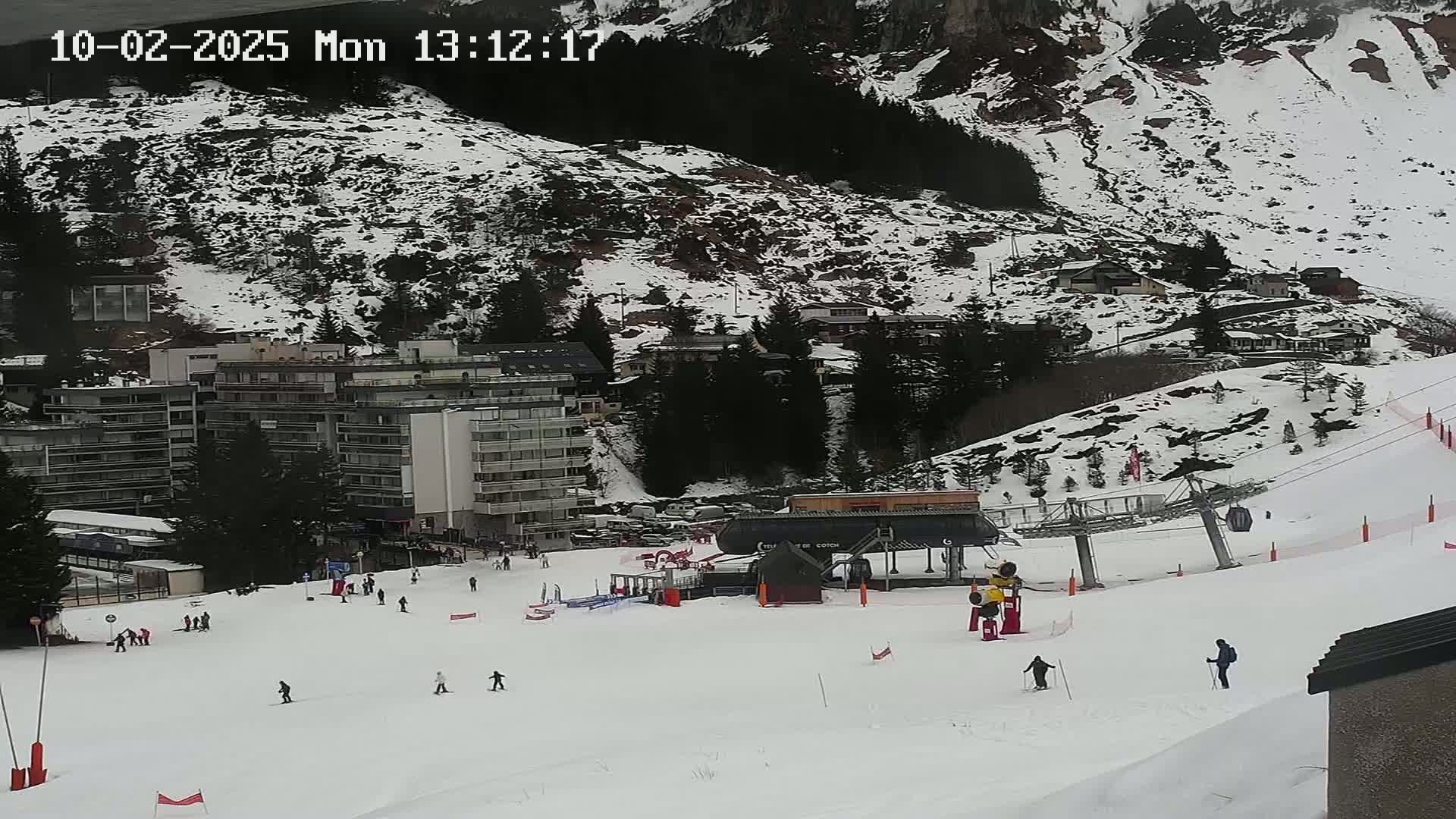 A ski slope with a chairlift and several skiers, situated below a snow-covered mountainside with buildings.
